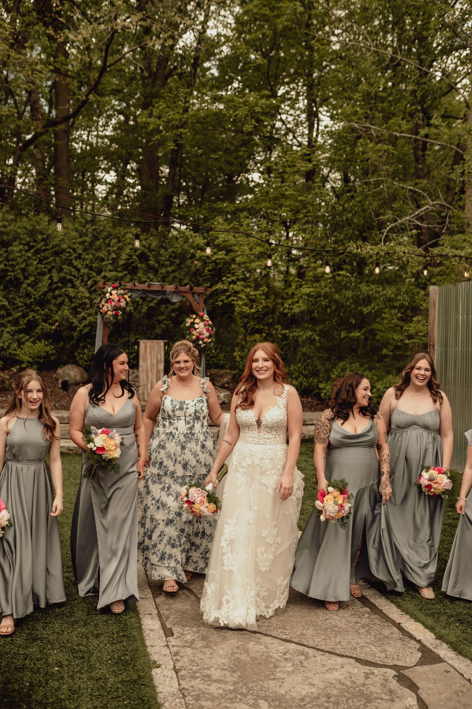 A bridal party in flowing gray dresses walks along a garden path under a floral arch during an outdoor wedding ceremony.