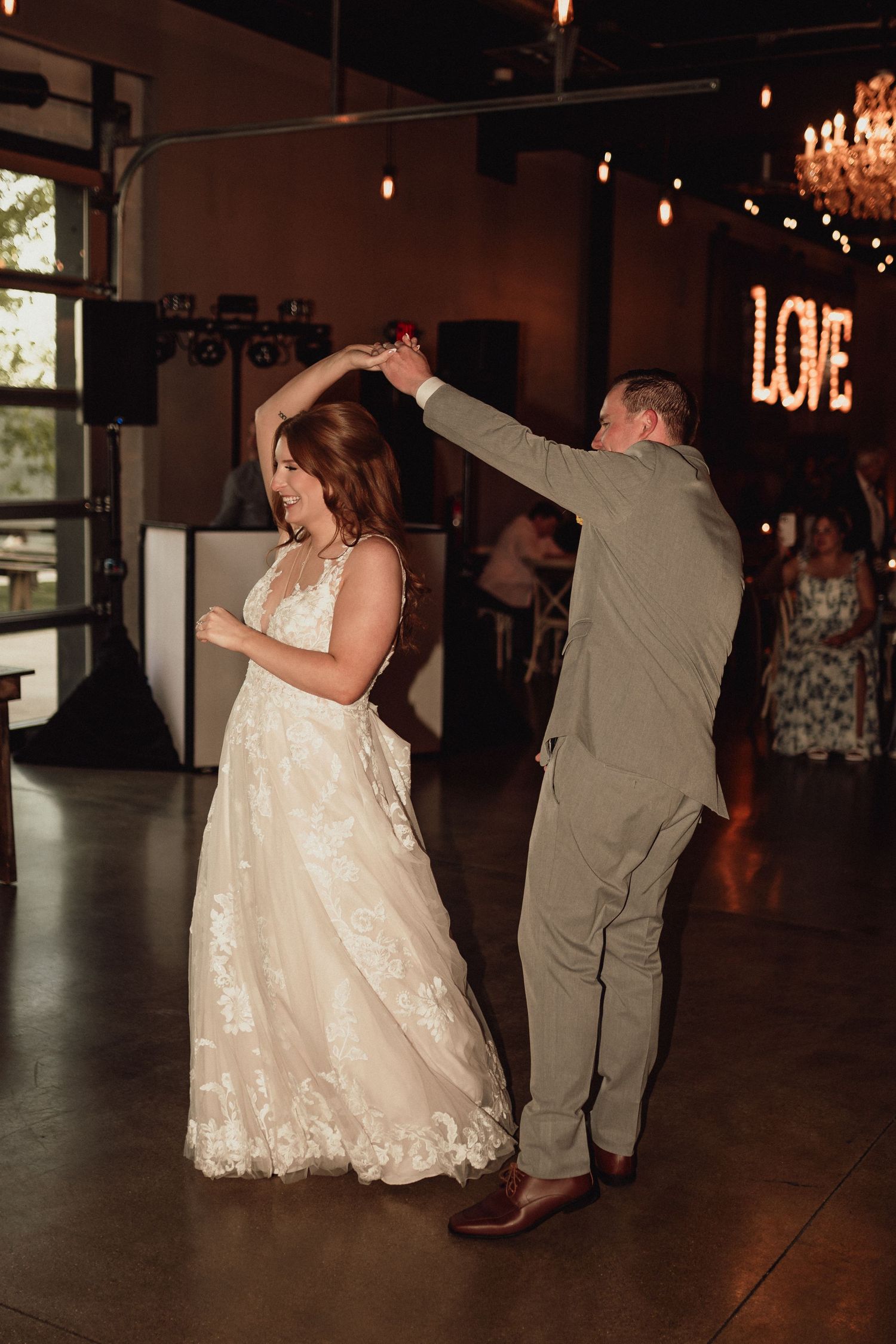 A bride in a lace dress dances with her partner in a dimly lit reception venue.