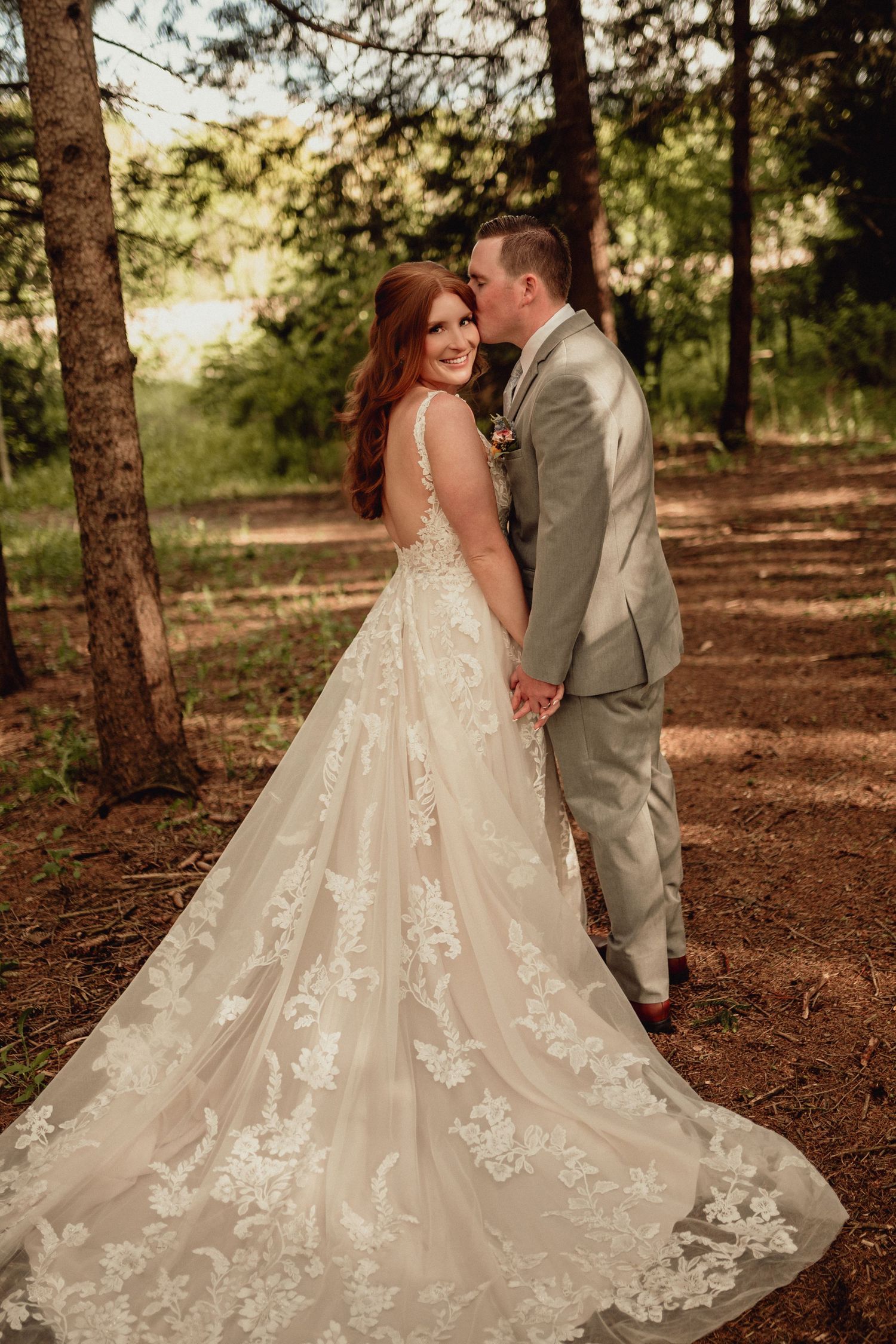 A romantic wedding couple embraces in a sunlit forest setting with a flowing lace wedding gown trailing behind them.