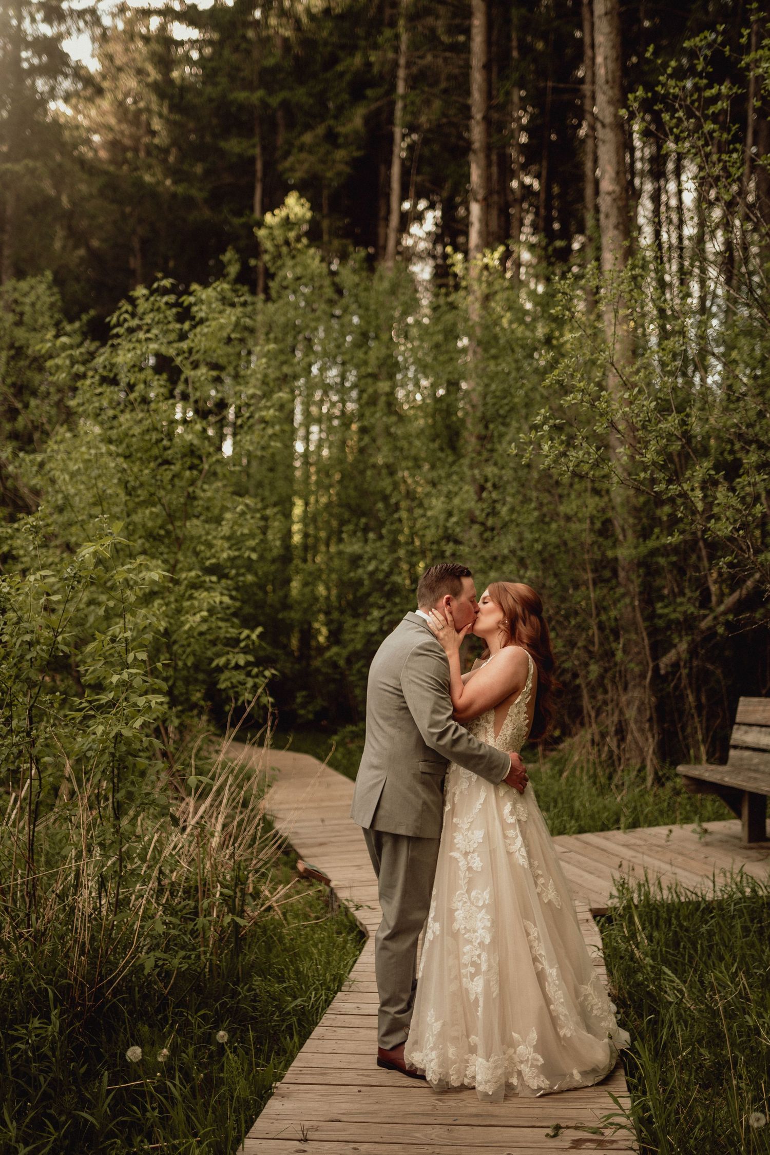 A couple shares a romantic kiss on a wooden boardwalk surrounded by lush green forest vegetation.