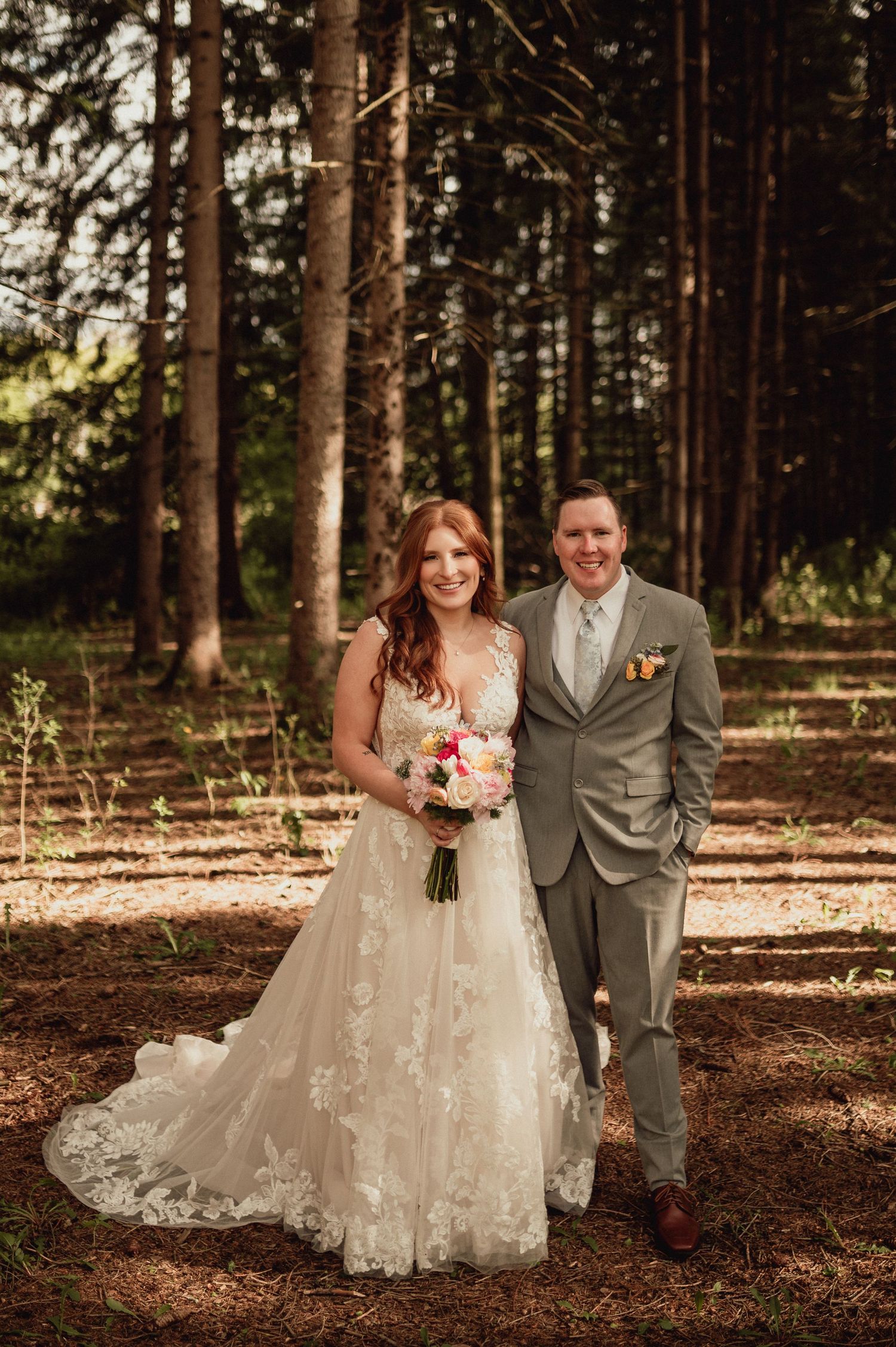 A bride in a lace wedding dress and groom in gray suit pose together in a sunlit forest setting.