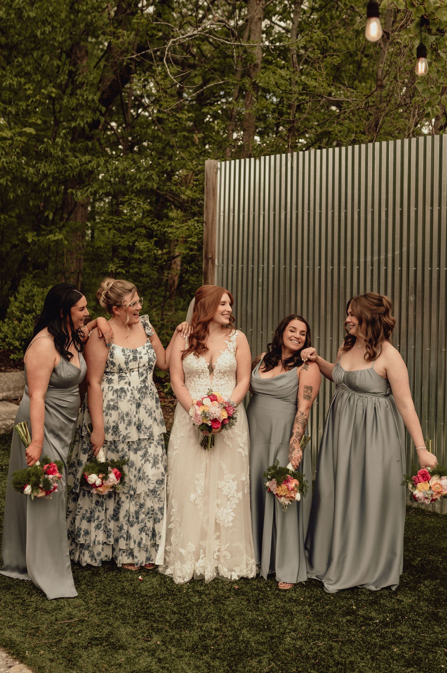 A bridal party in gray dresses and floral patterns poses together on a lawn in front of a wooden fence.