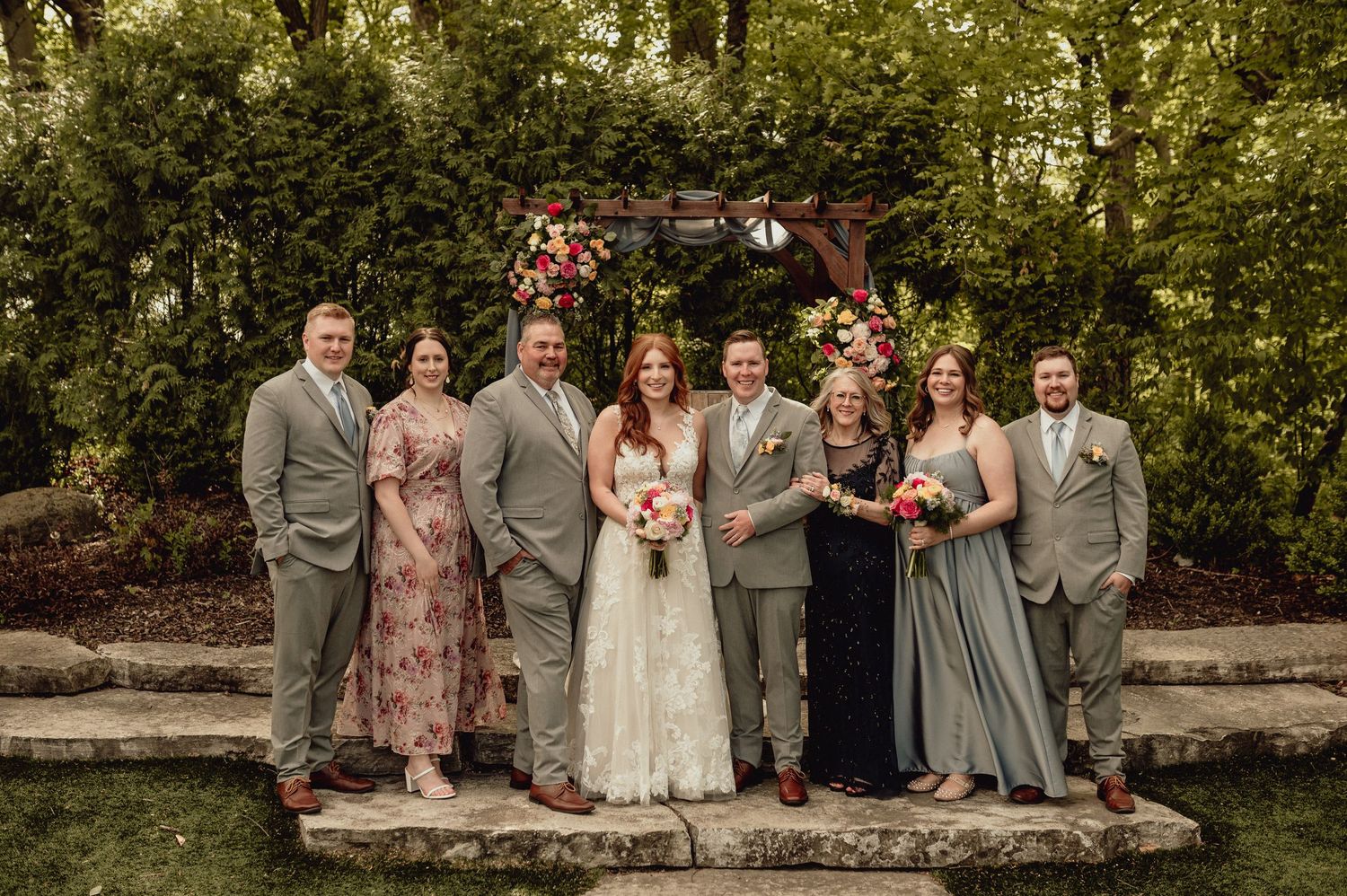 Wedding party in gray and pink attire poses together on stone wall in outdoor garden setting.