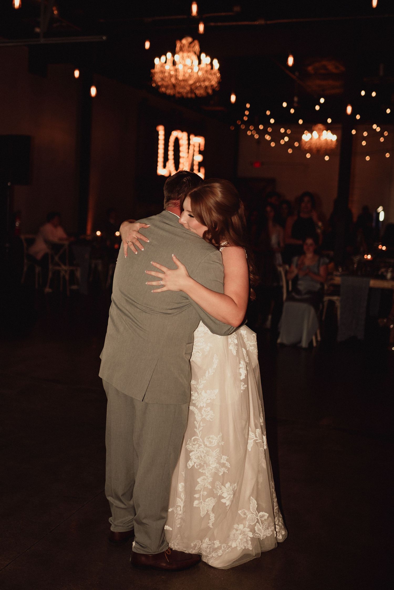 A couple shares an intimate dance moment at a wedding reception under warm lighting and chandeliers.