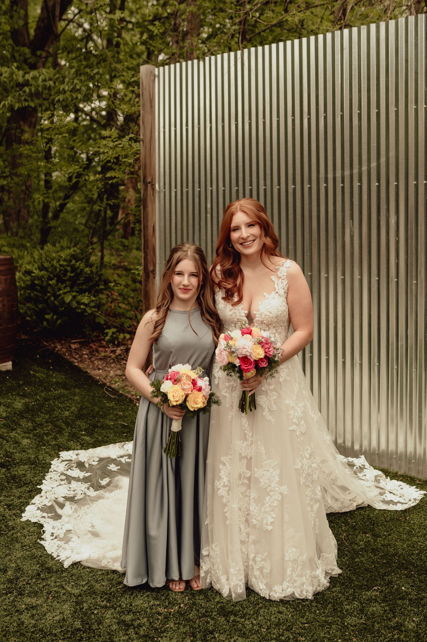 Wedding party members stand together holding pink and peach bouquets in front of industrial metal wall.