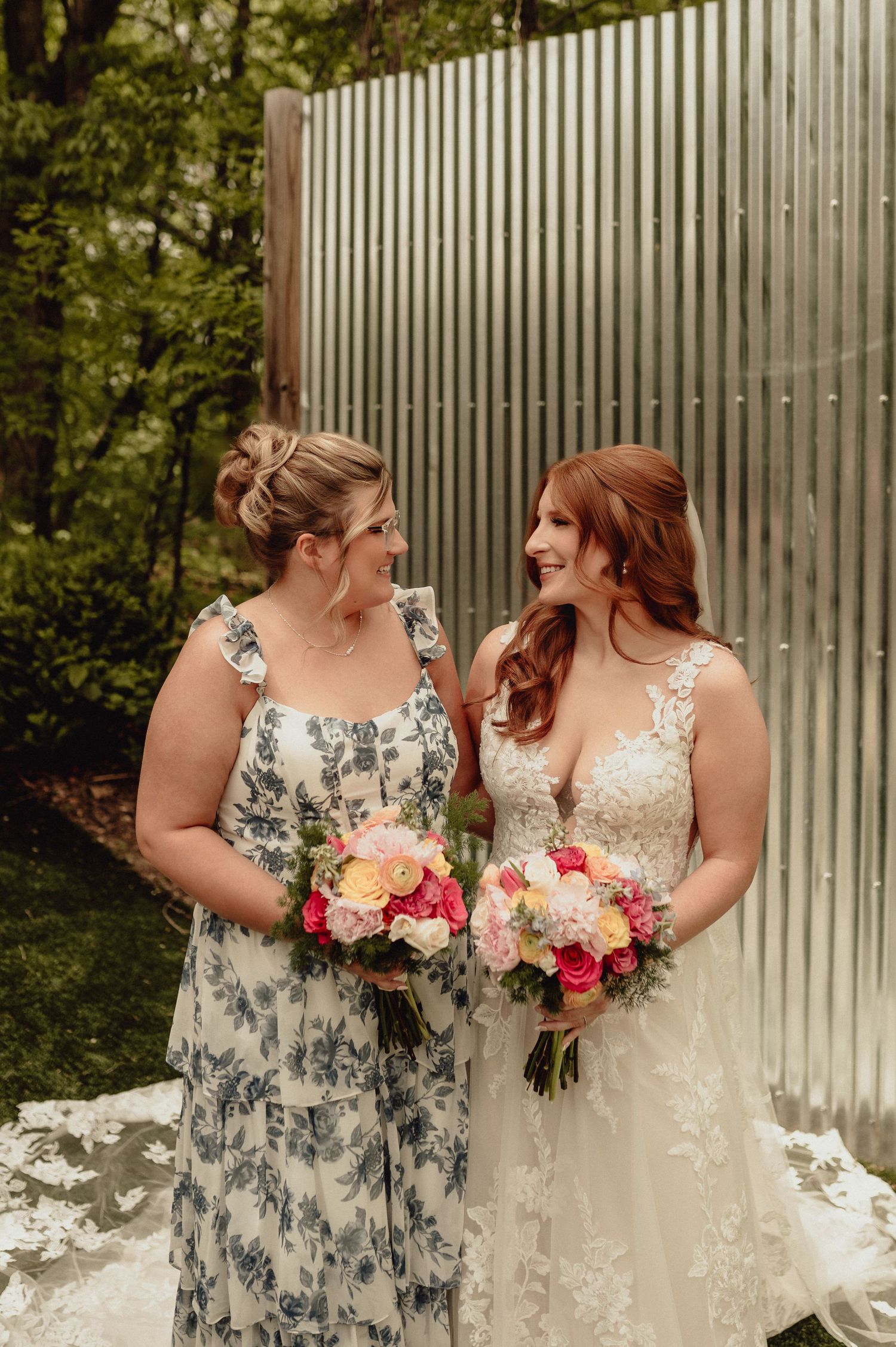 Two people in wedding attire share a joyful moment together while holding colorful floral bouquets against a white fence.