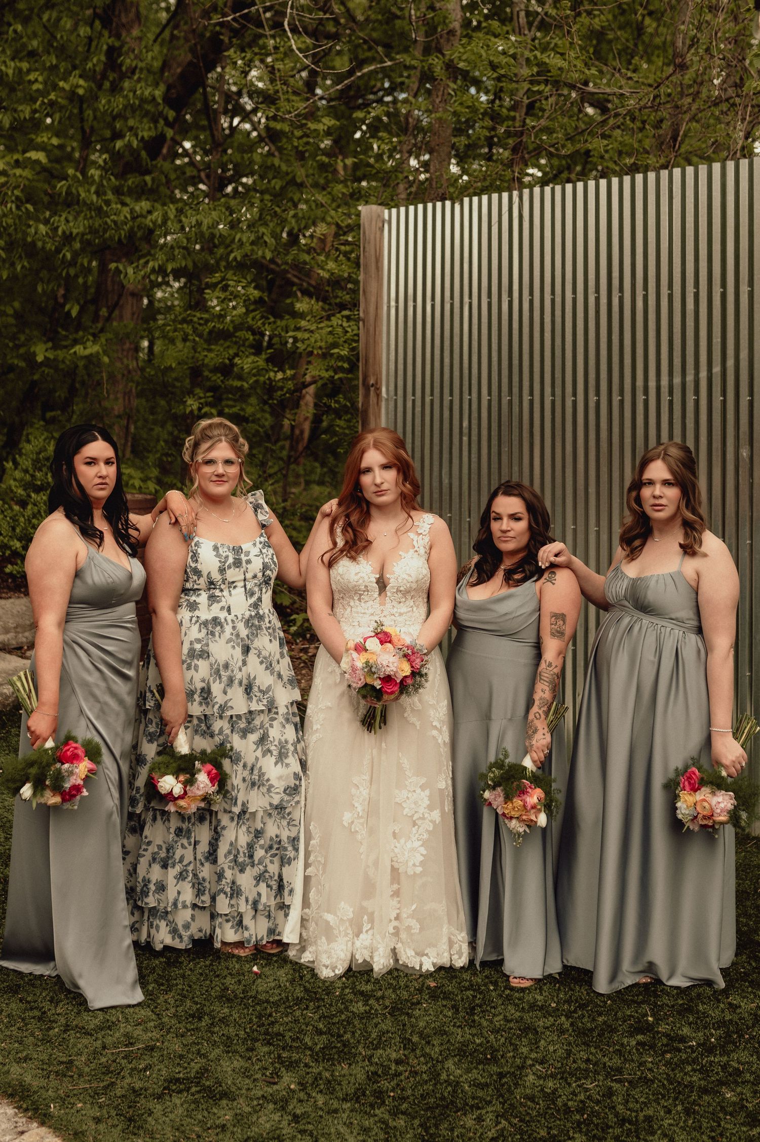 Bridesmaids in sage green dresses pose with bride in white lace gown holding pink and peach bouquets in garden setting.