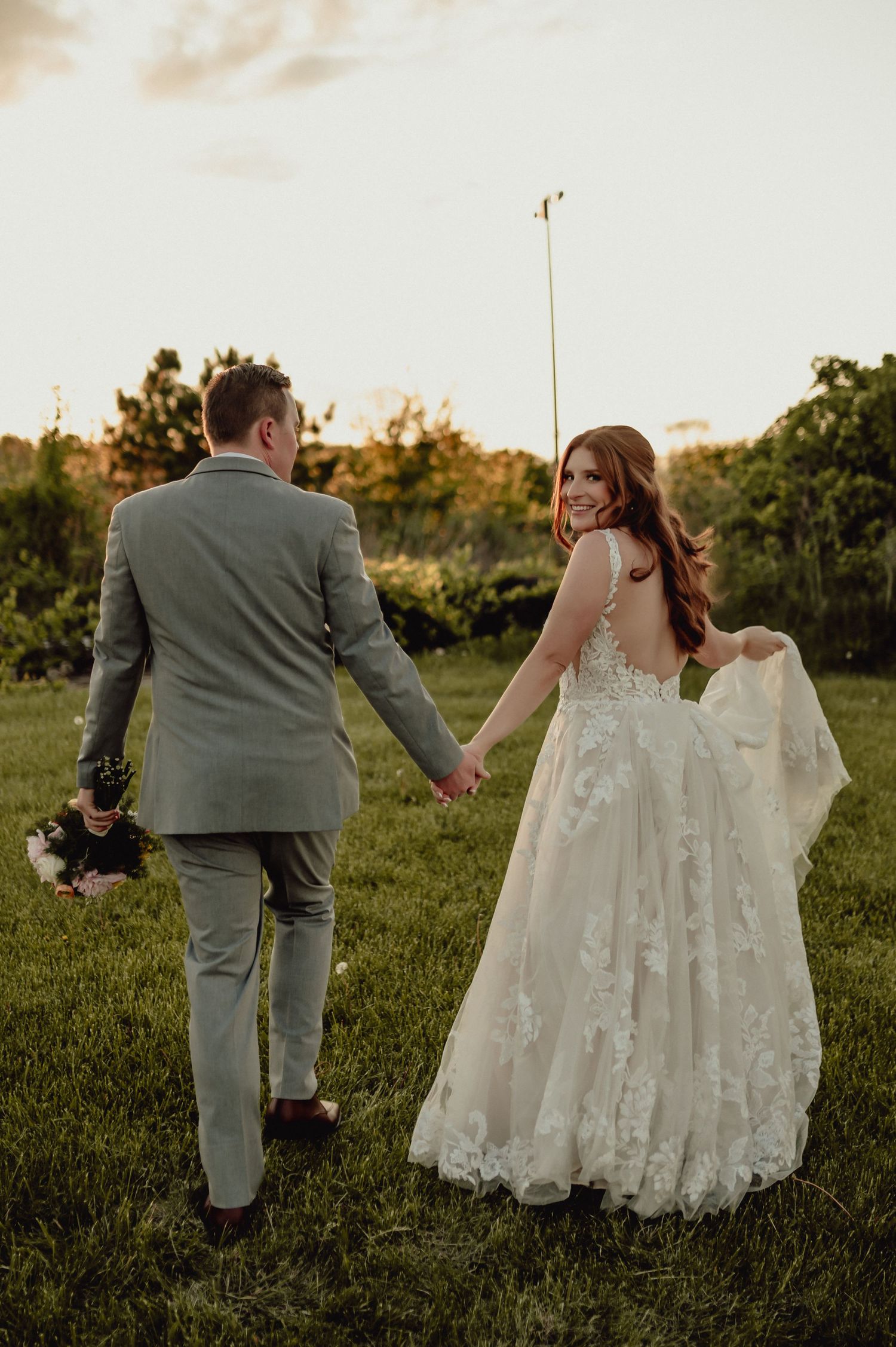 A couple in wedding attire walk hand in hand through a sunlit grassy field during golden hour.