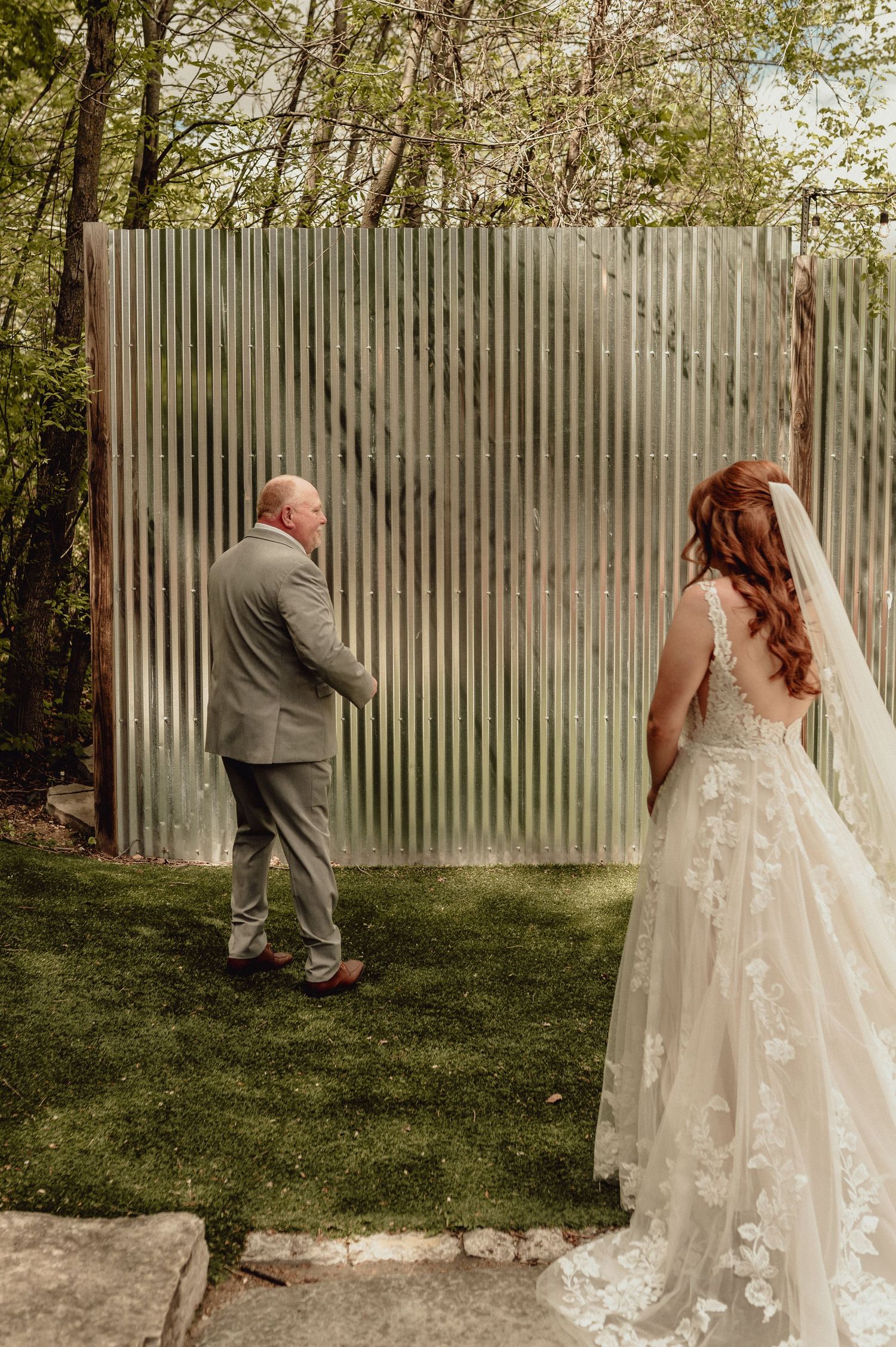 A wedding couple plays hide and seek around a wooden fence in a backyard garden setting.