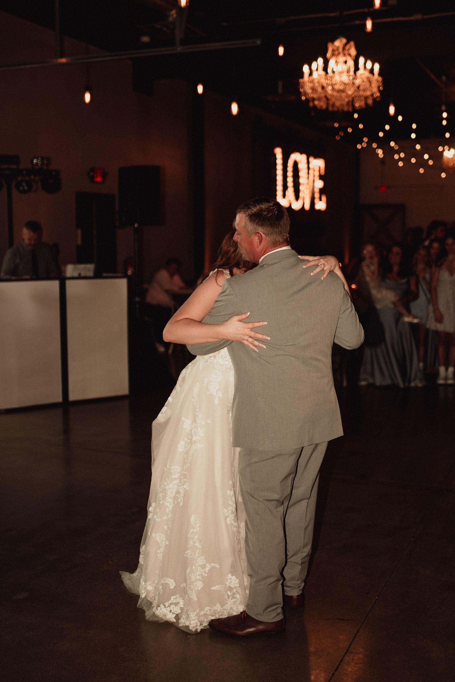 A couple dances together in a dimly lit reception hall with a crystal chandelier overhead.