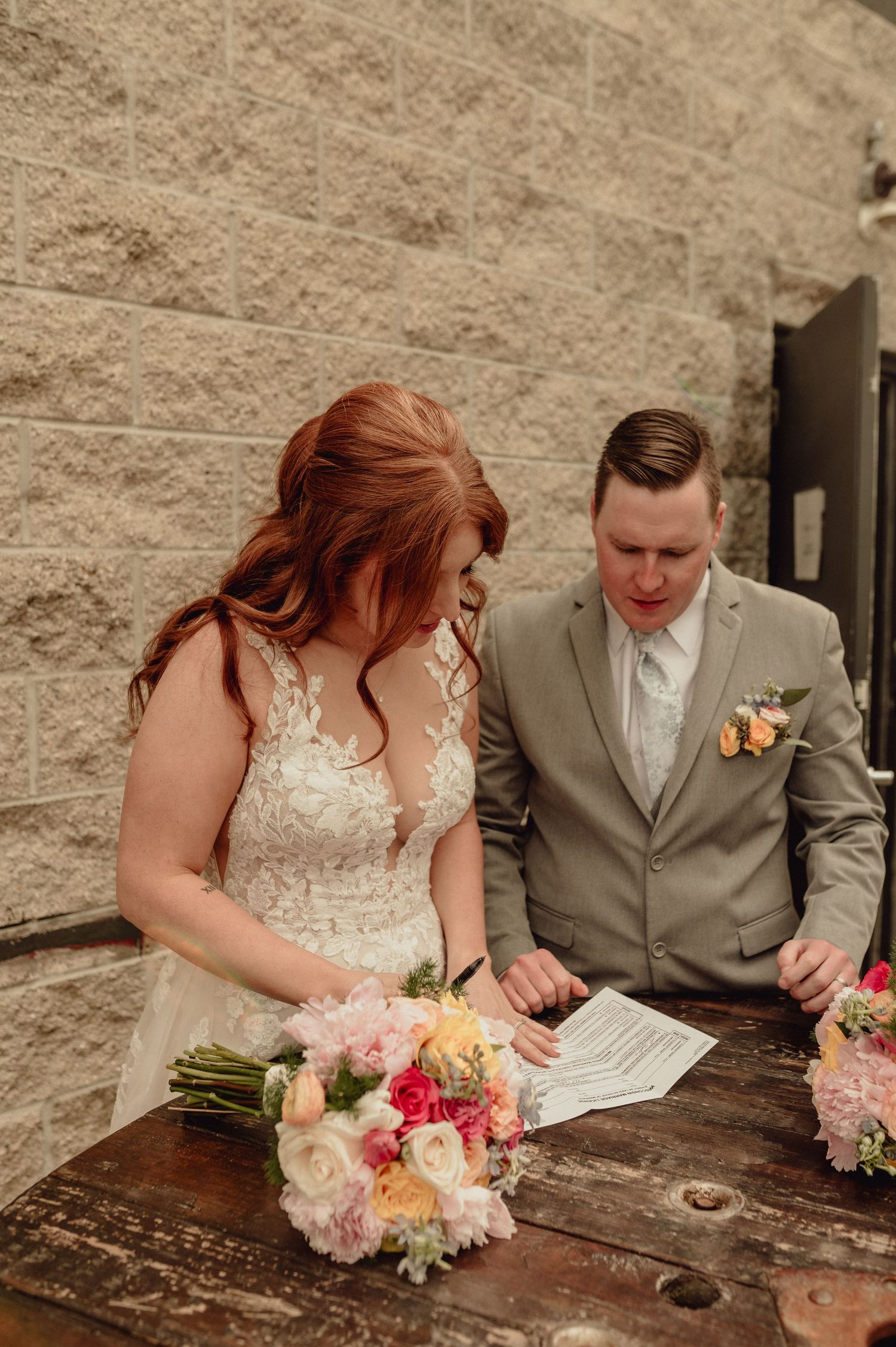 A couple in wedding attire signs documents together at a rustic wooden table with pink and peach floral bouquet nearby.