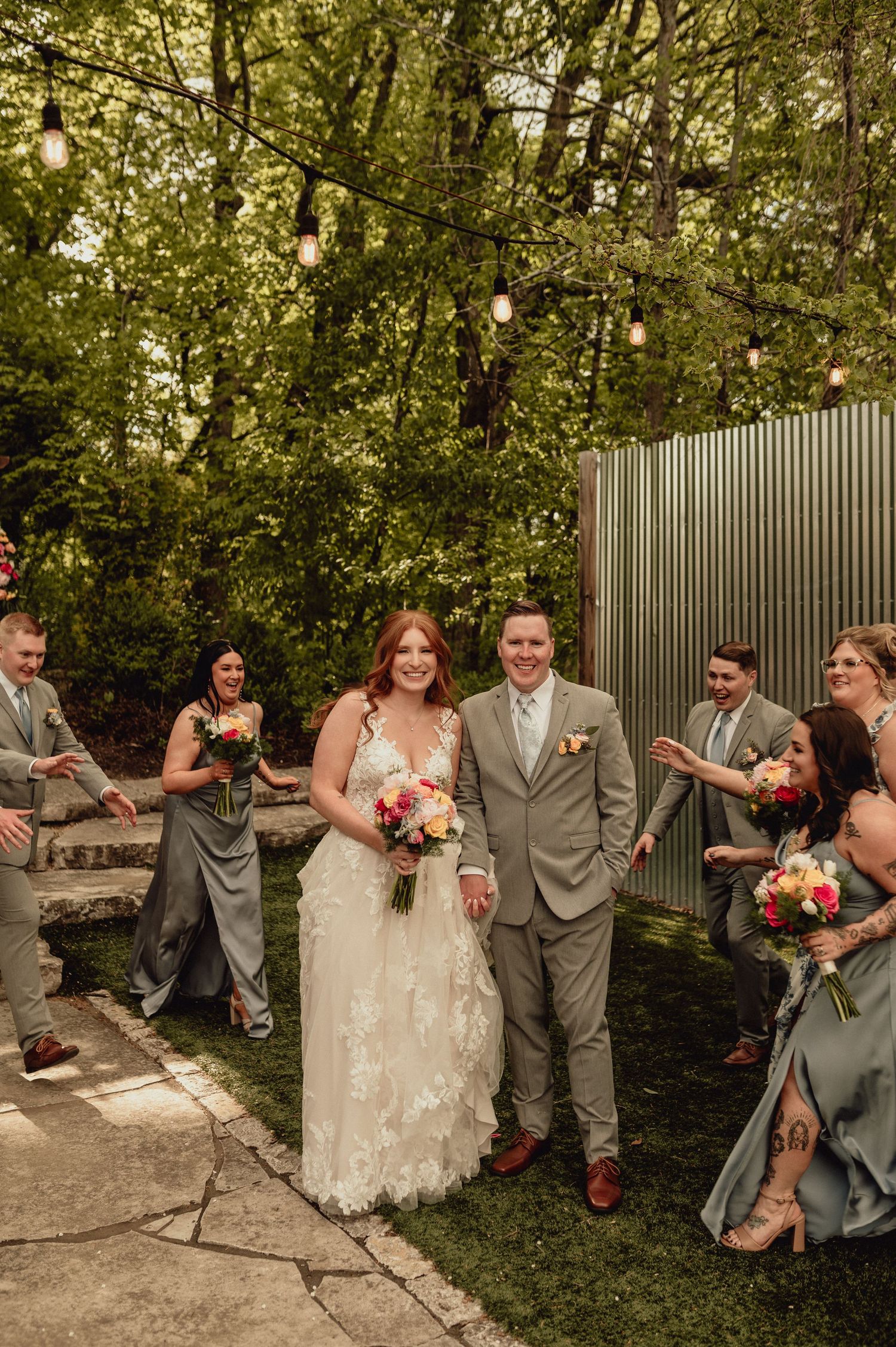 Wedding party poses playfully in a garden setting with lush greenery and a wooden fence backdrop.