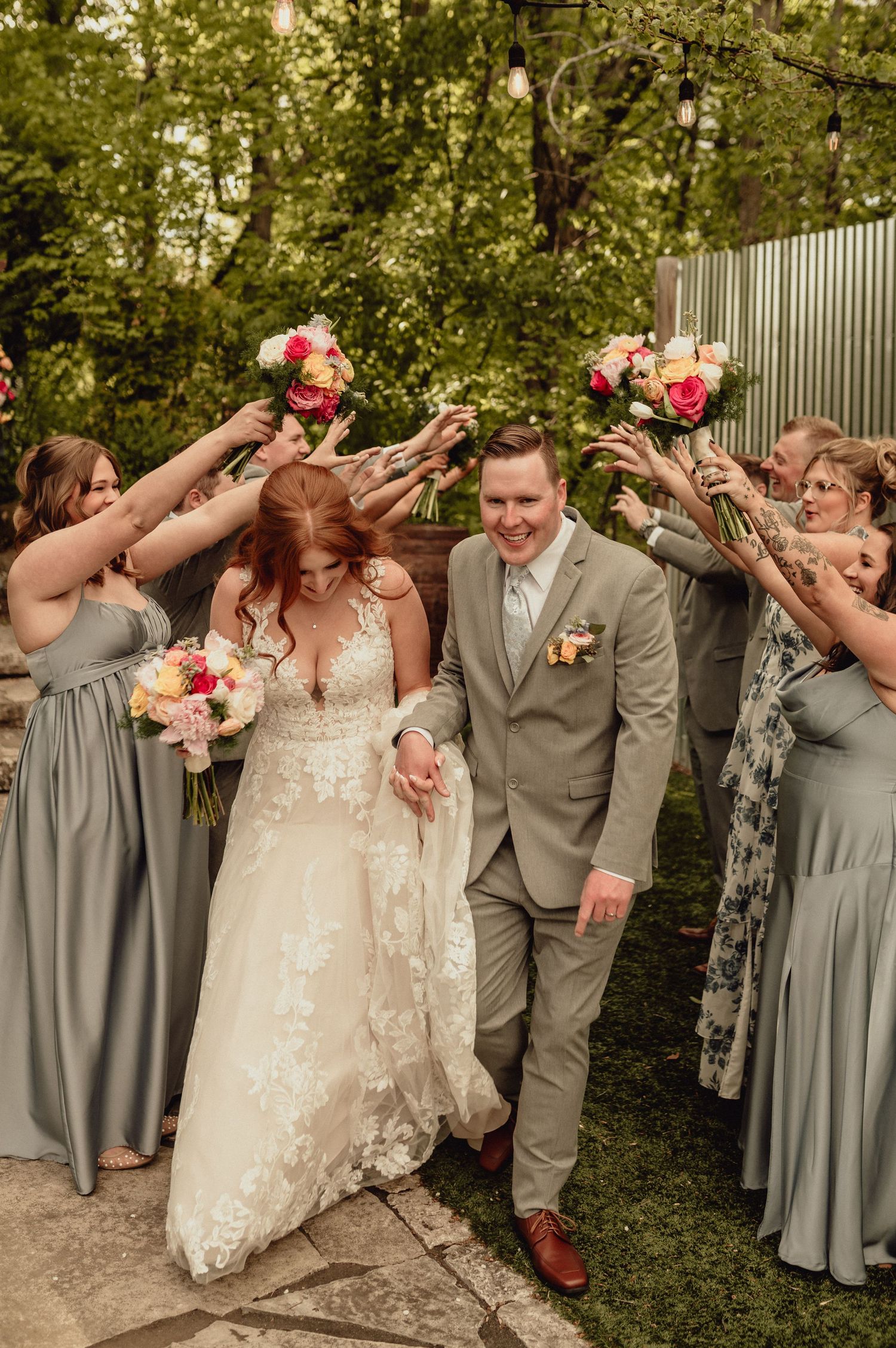 Wedding party in gray attire celebrates joyfully in an outdoor garden setting with pink floral bouquets.