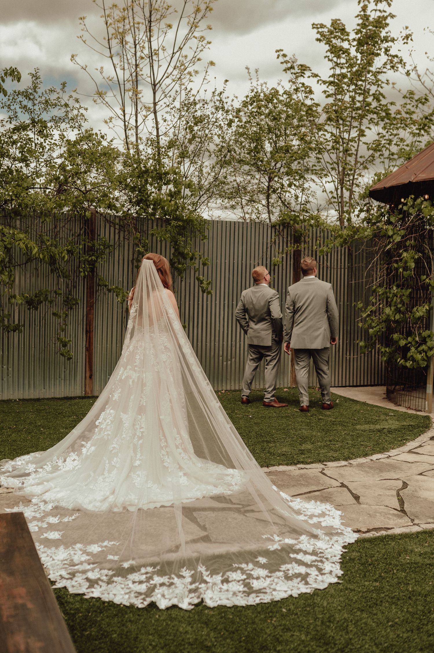 A wedding dress with an ornate lace train spreads across green grass while two people in gray suits stand in the distance.