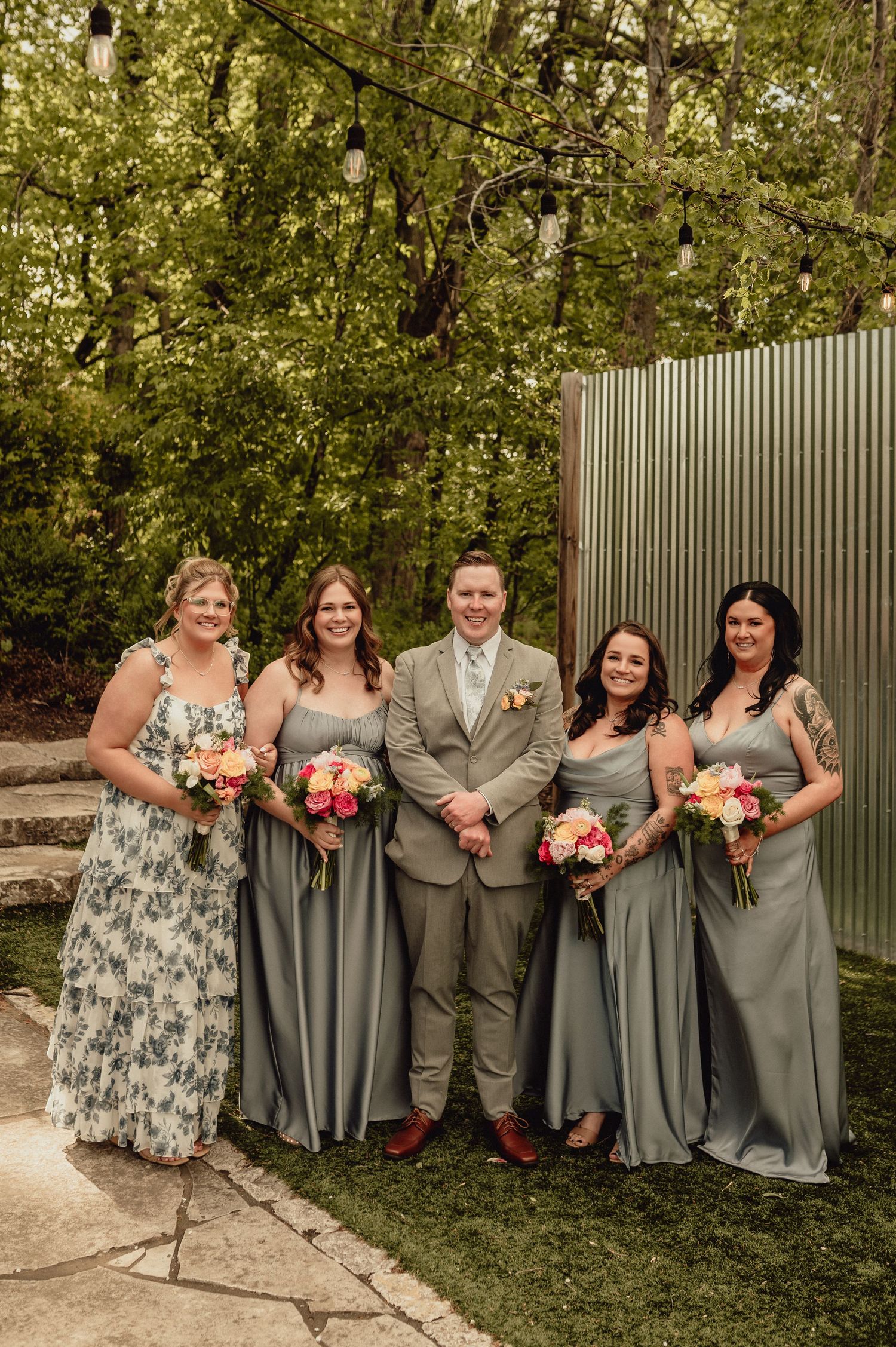 Wedding party in gray dresses and suits poses together against green foliage backdrop.