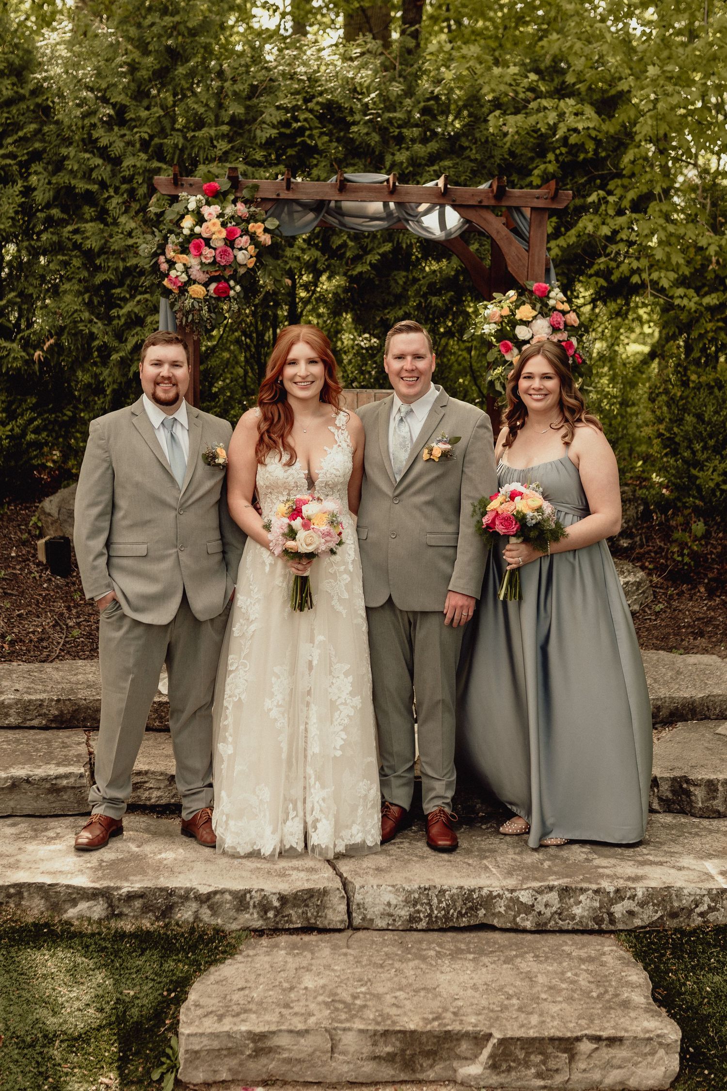 Wedding party in gray attire and white dress poses together on stone wall in garden setting.