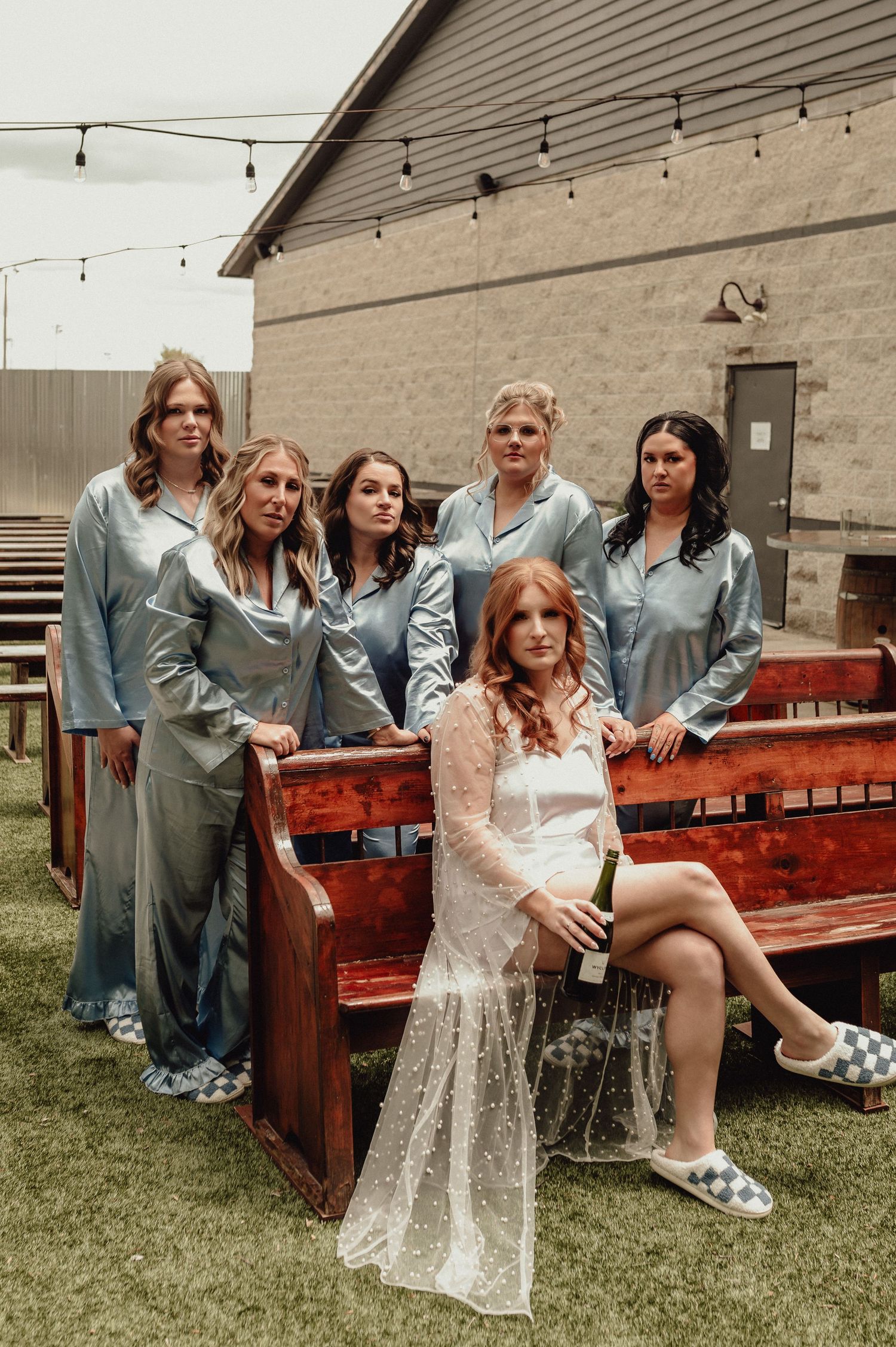 A group in coordinated blue outfits and one in white lace pose together on a rustic wooden bench outdoors.