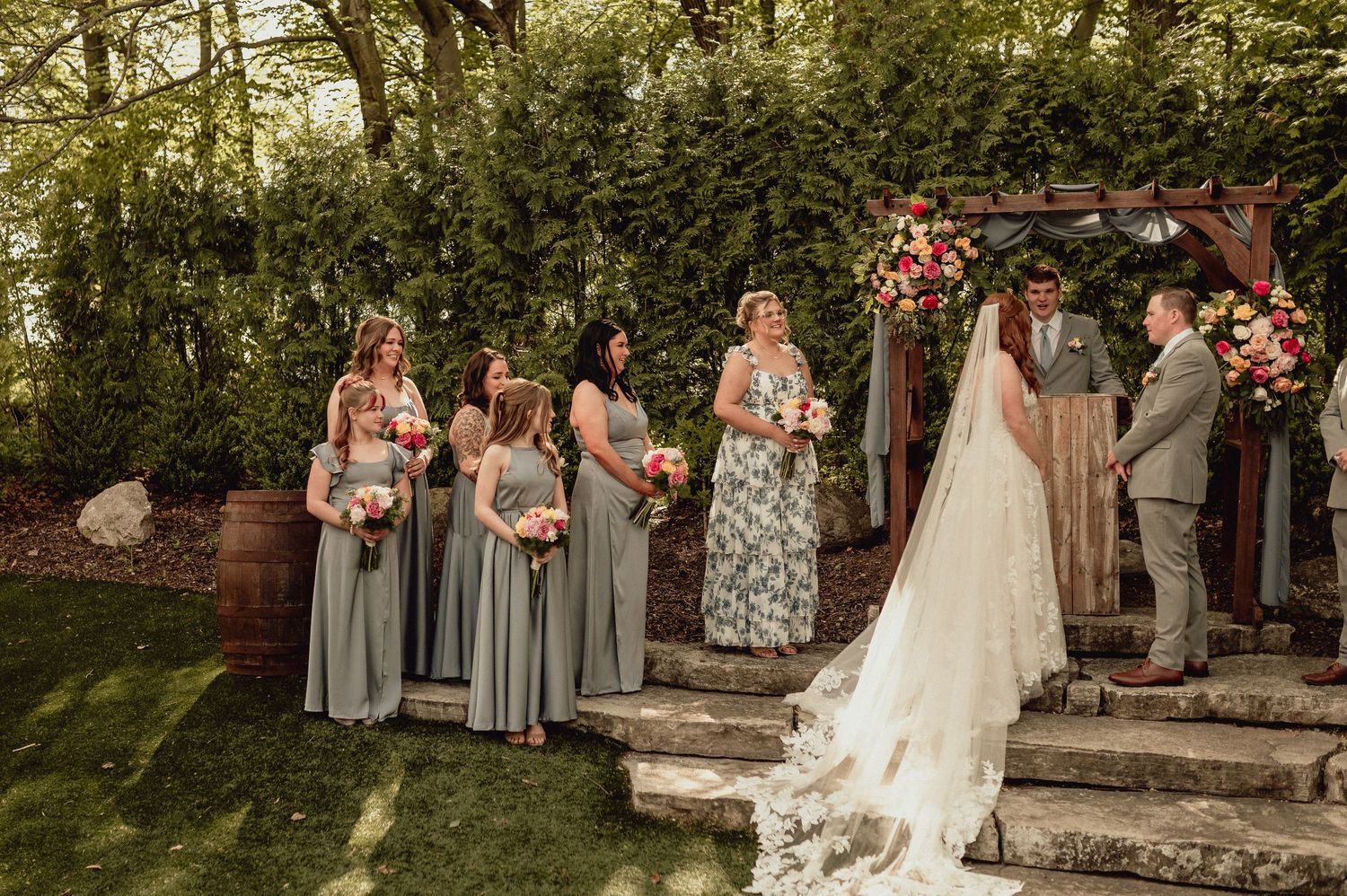 A wedding party poses together in a garden setting with lush greenery and floral arrangements.