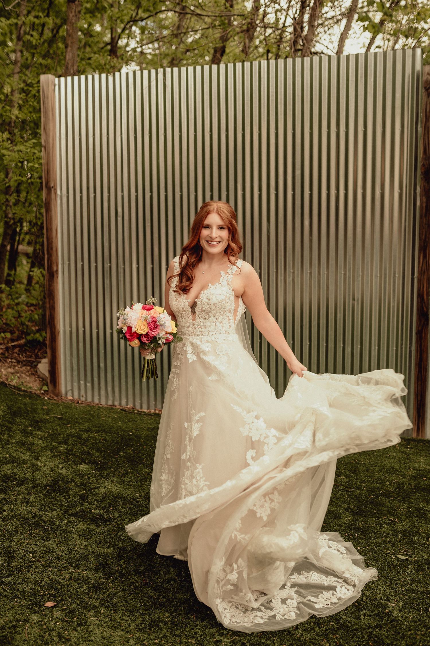 A bride in a flowing lace wedding gown poses against a corrugated metal backdrop in an outdoor garden setting.