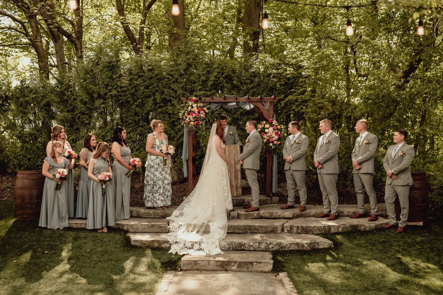 Wedding party poses outdoors in a garden setting with stone steps and lush greenery creating an elegant backdrop.