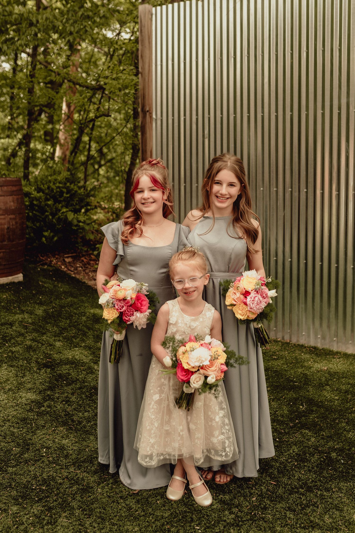 Bridesmaids in gray dresses and flower girl in lace dress pose with coral and pink bouquets in garden setting.