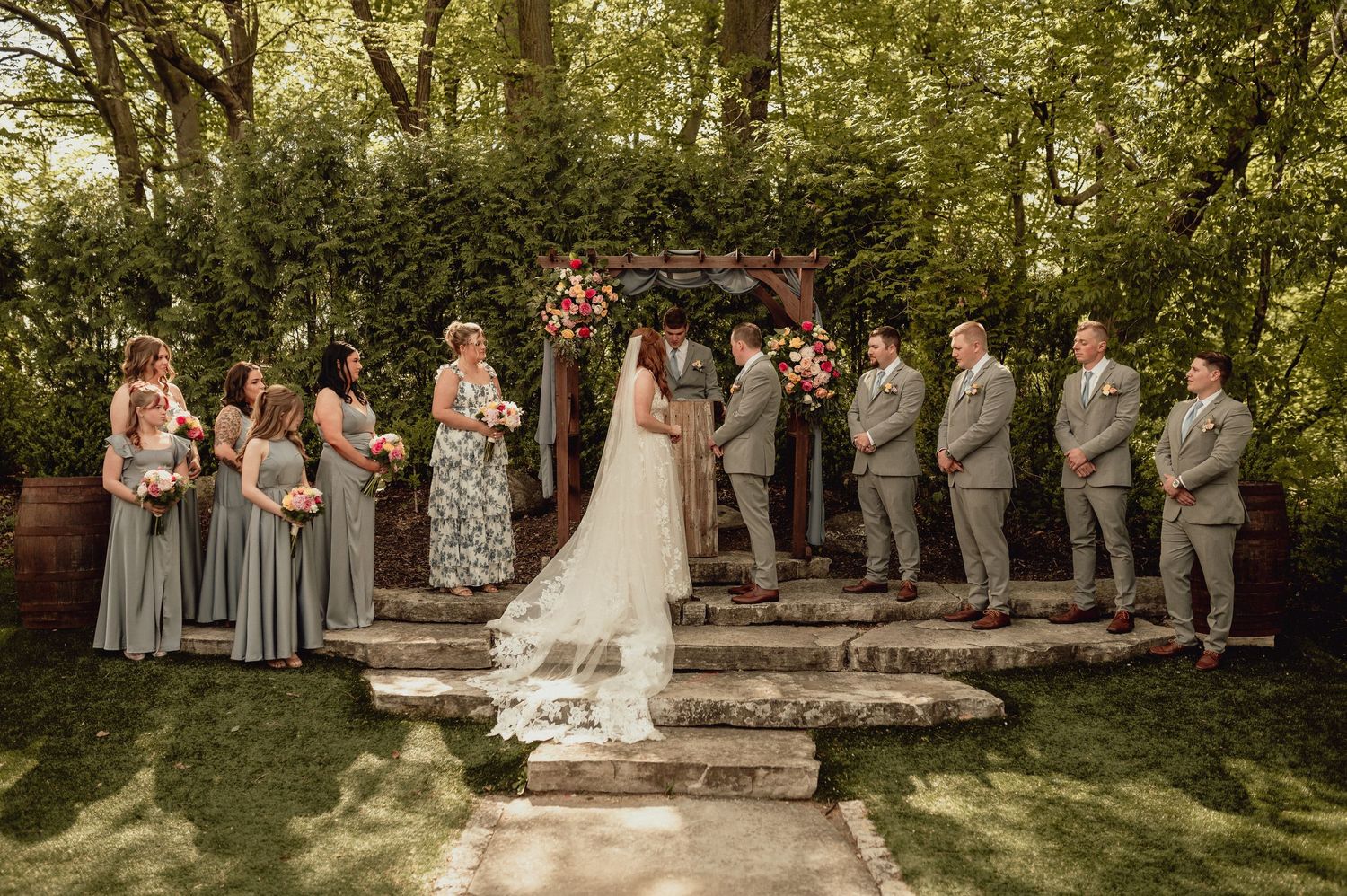 Wedding party stands in a line on stone steps surrounded by lush greenery in an outdoor garden setting.