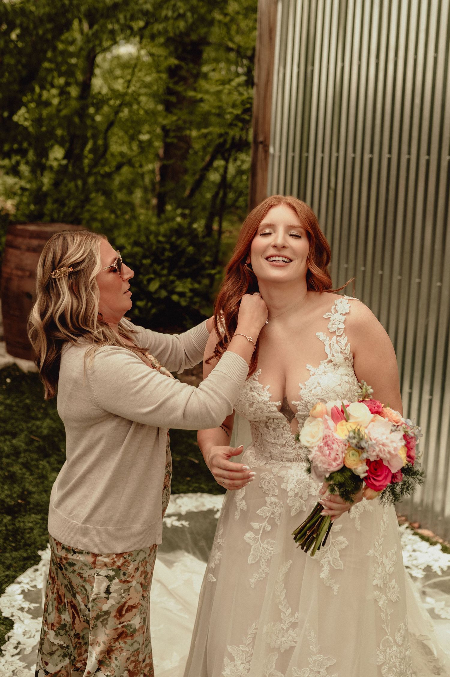 Two women share a joyful moment in a garden setting, one wearing a wedding dress and holding a colorful bouquet.
