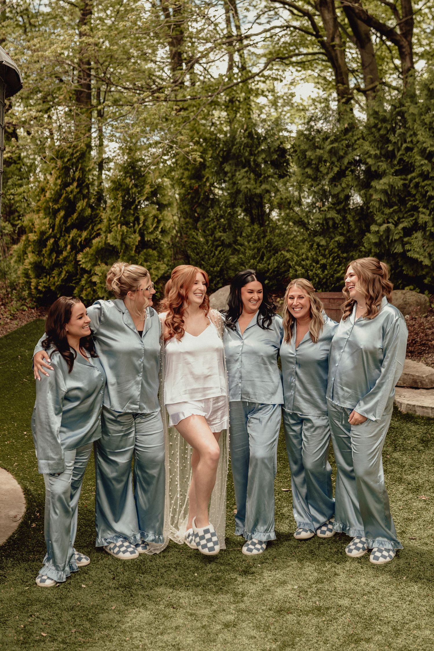 Wedding party wearing matching silk robes and slippers pose together playfully in a garden setting.