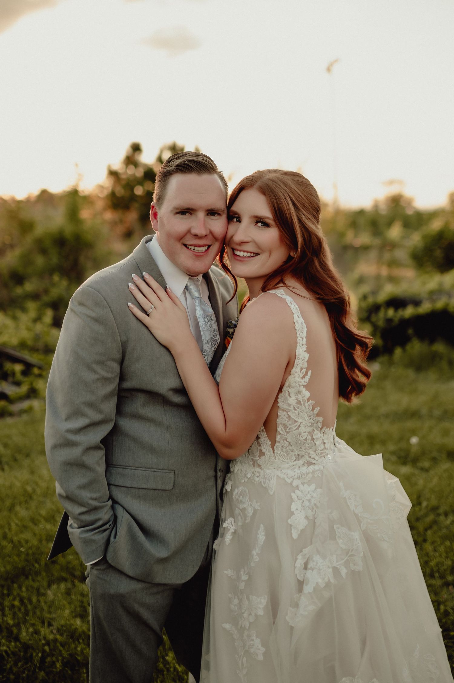 A romantic wedding couple embraces in a sunlit field while wearing a grey suit and lace wedding gown.