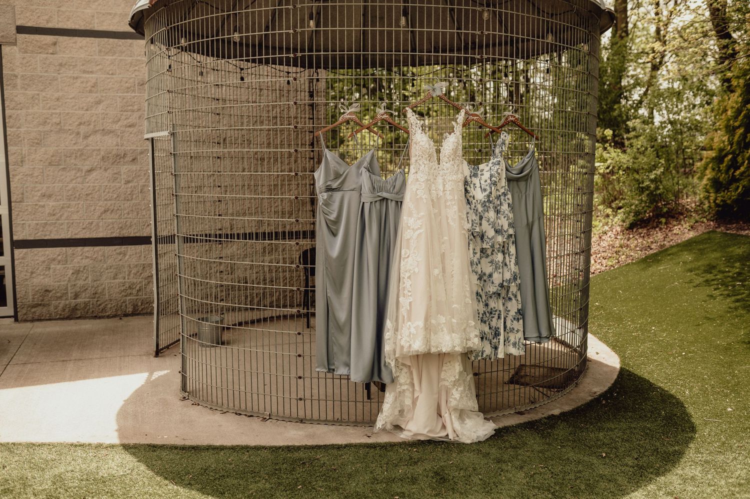 Vintage wedding dress displayed on a hanger next to a metal bird cage in an outdoor garden setting.
