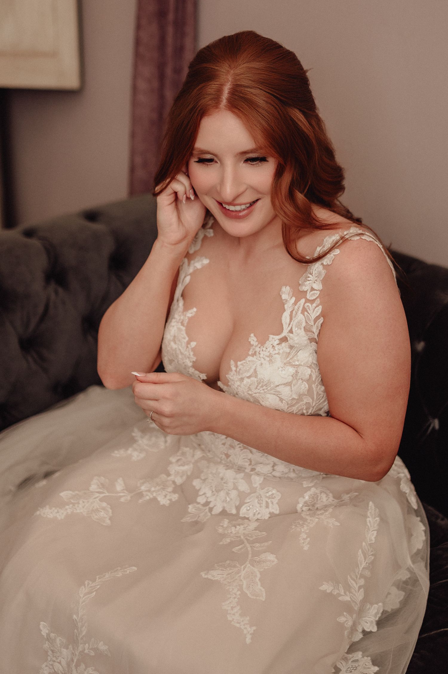 Bride in a white lace wedding dress with floral appliques laughs joyfully while sitting on a dark couch.