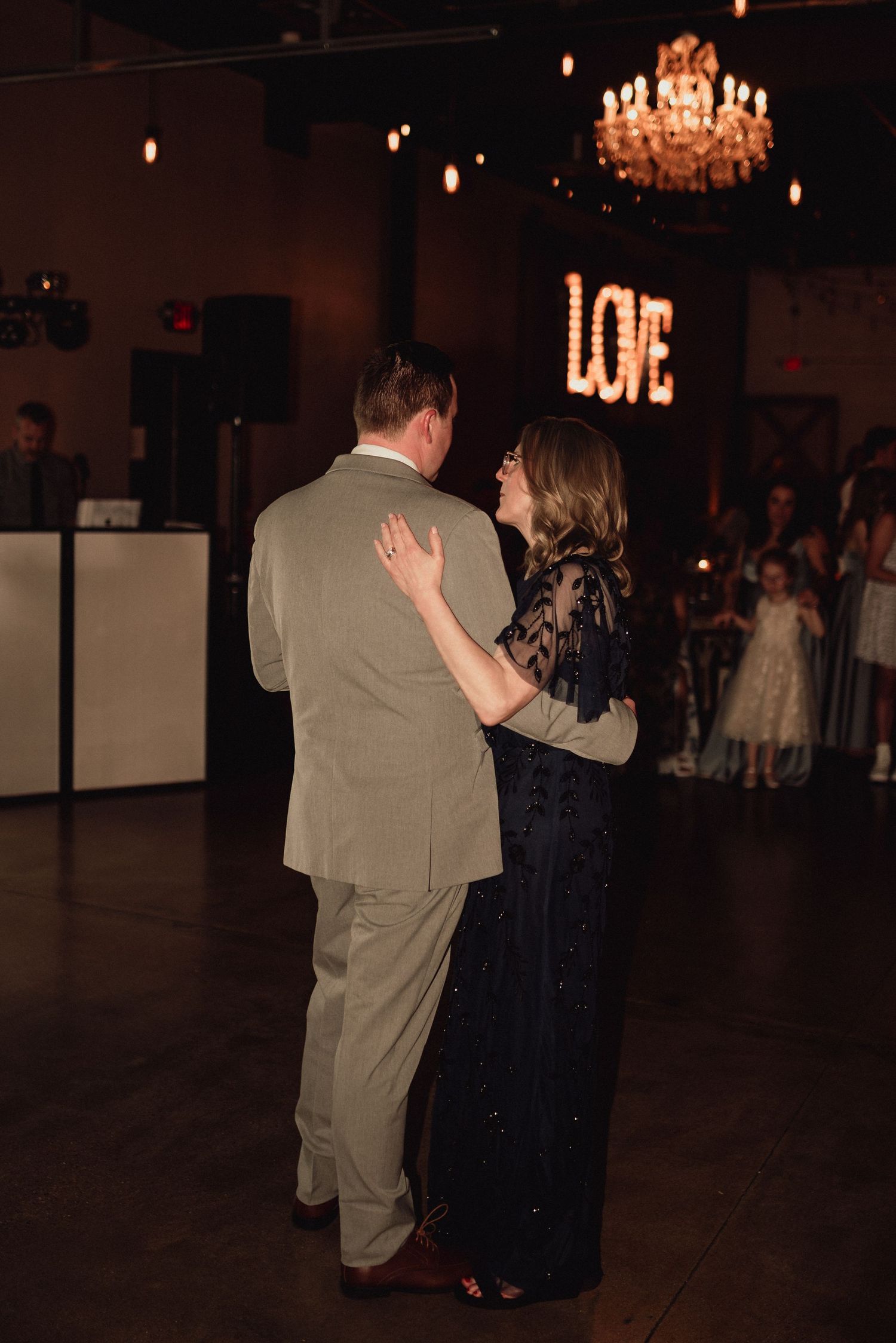 A couple shares a dance under a glowing LOVE sign and crystal chandelier at an elegant reception venue.