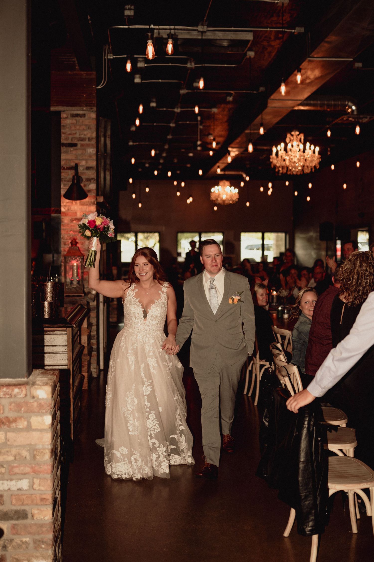Wedding ceremony procession in a rustic brick venue with chandeliers and string lights creating a warm ambiance.