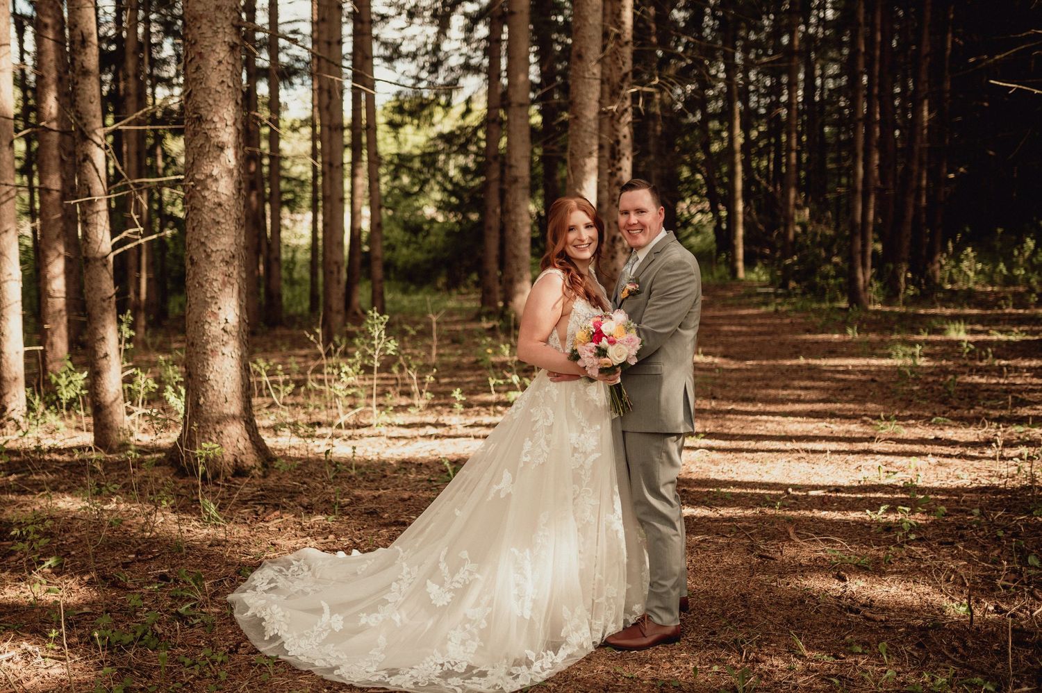 A couple embraces in a romantic forest setting with sunlight filtering through tall pine trees.