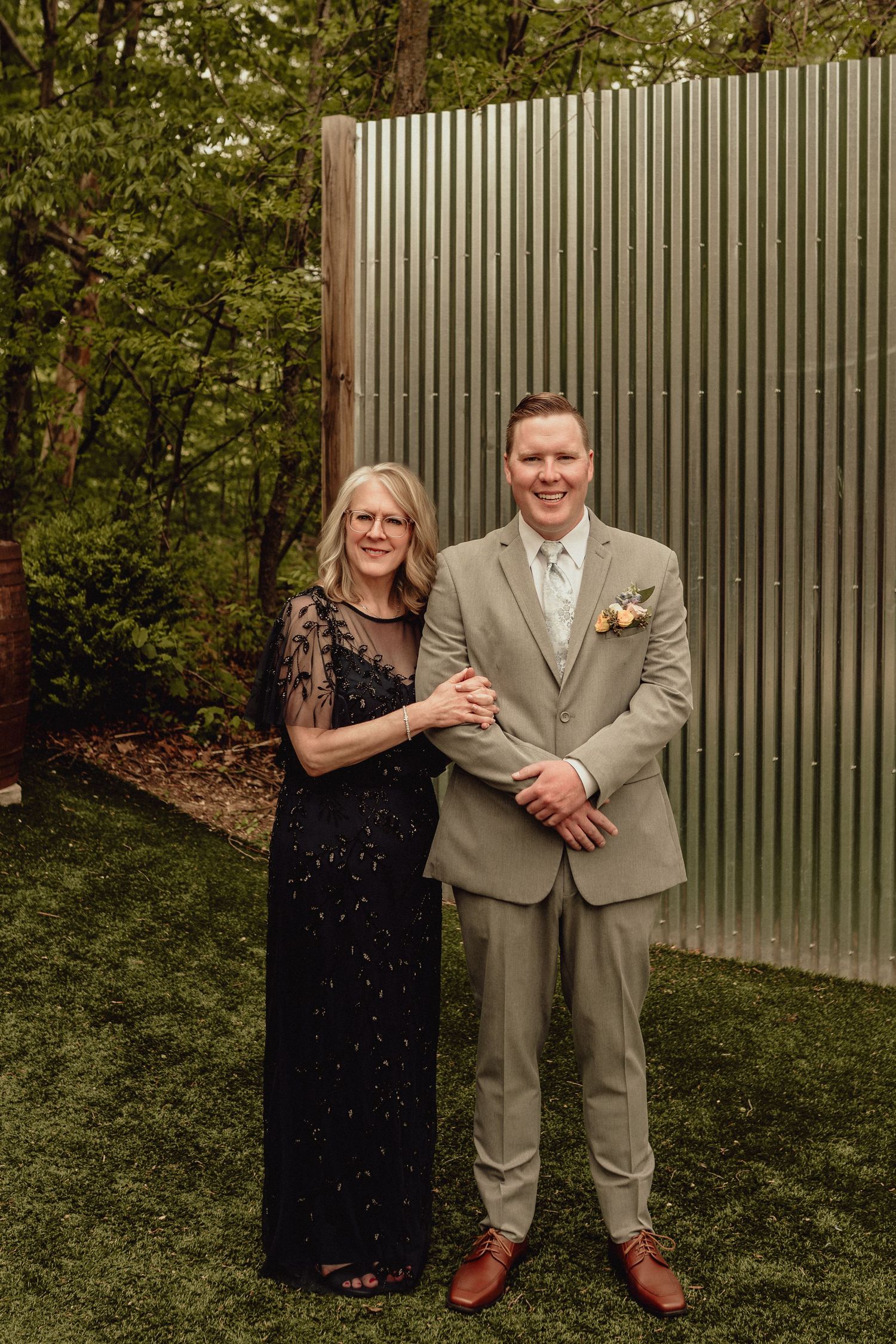 A couple shares playful moments in a garden setting with a grey fence backdrop.
