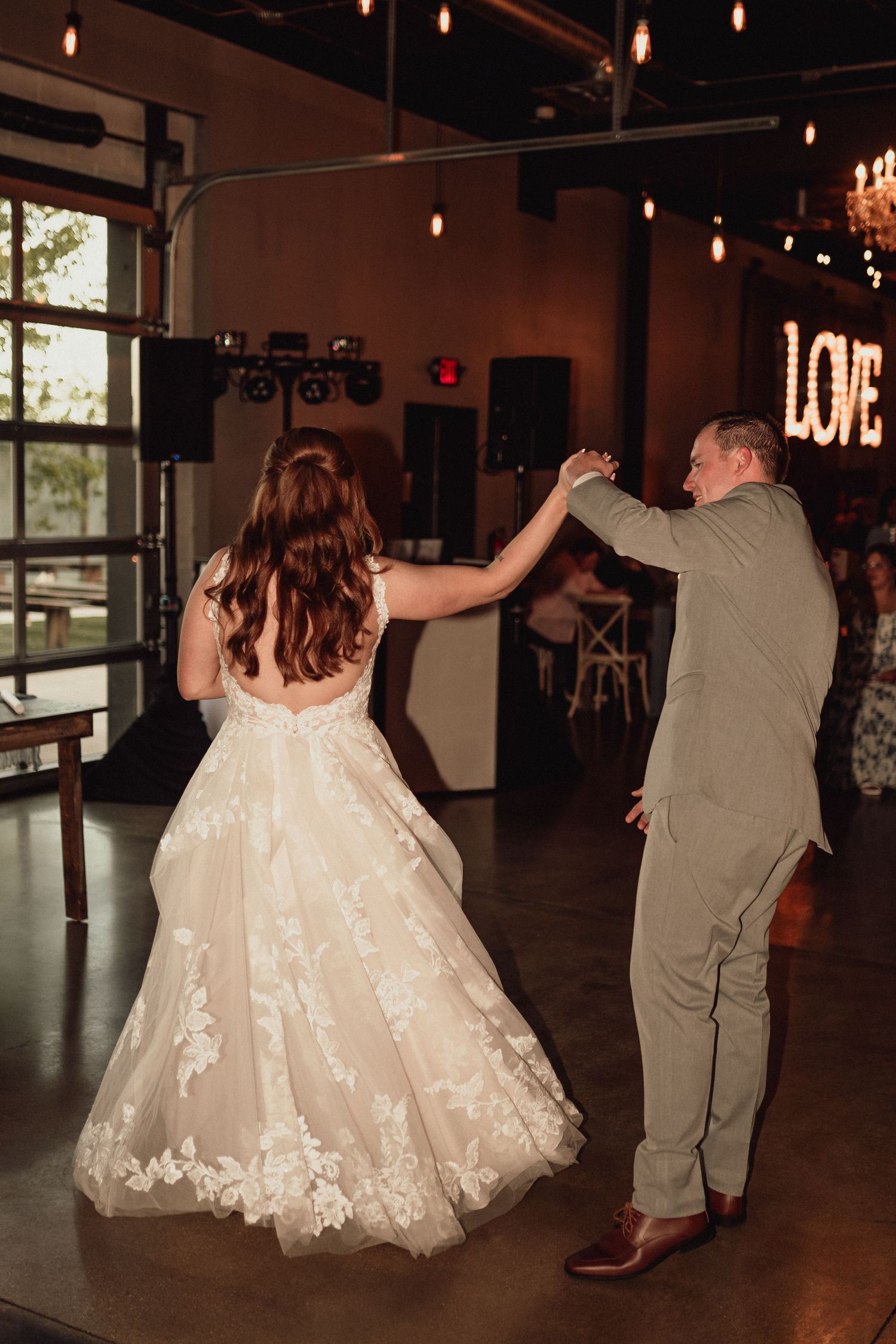 A couple dances at their wedding reception in a rustic venue with a glowing LOVE sign and pendant lights.