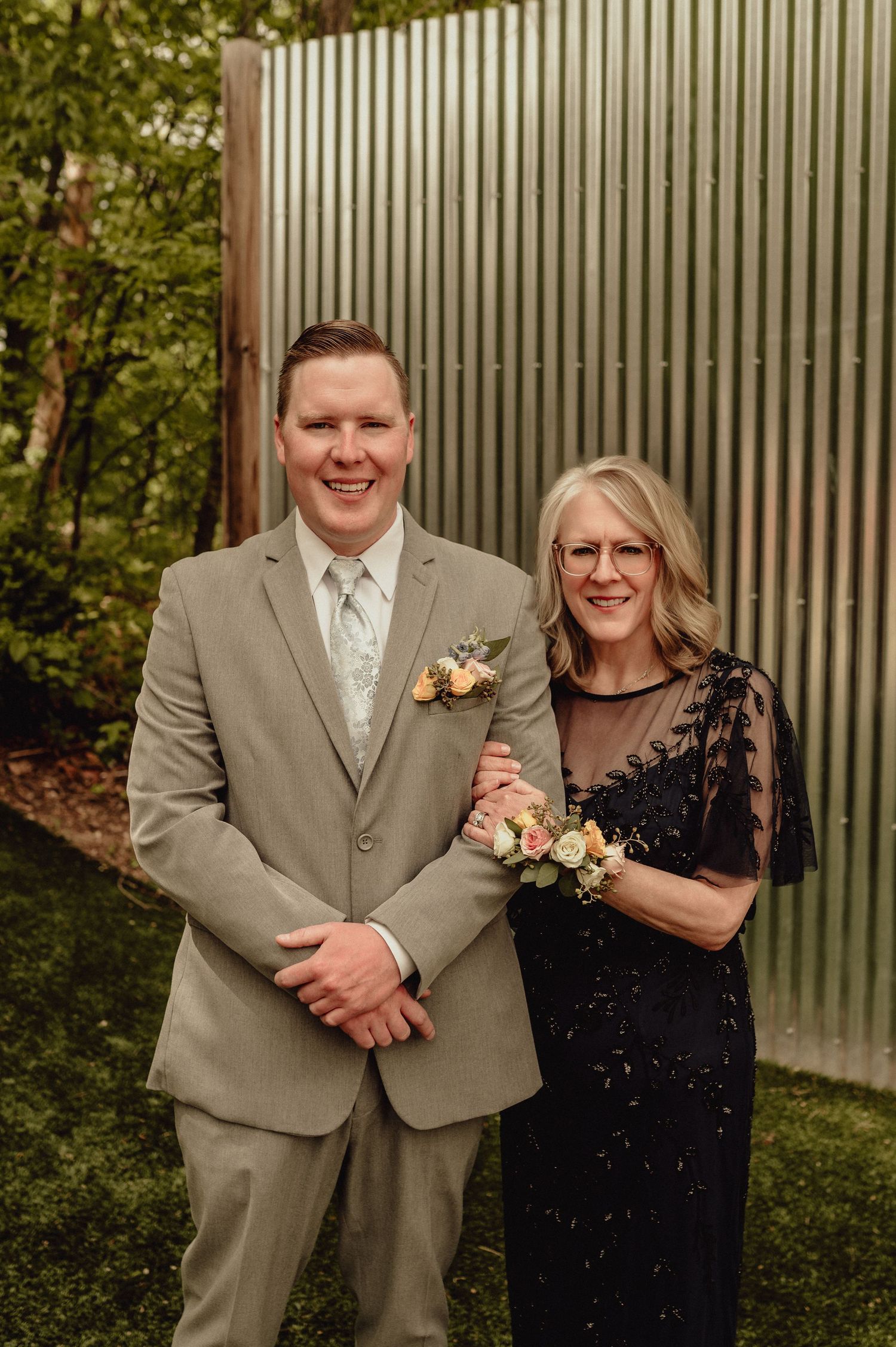 Formal portrait of two people in elegant attire against a metal wall backdrop at an outdoor event.