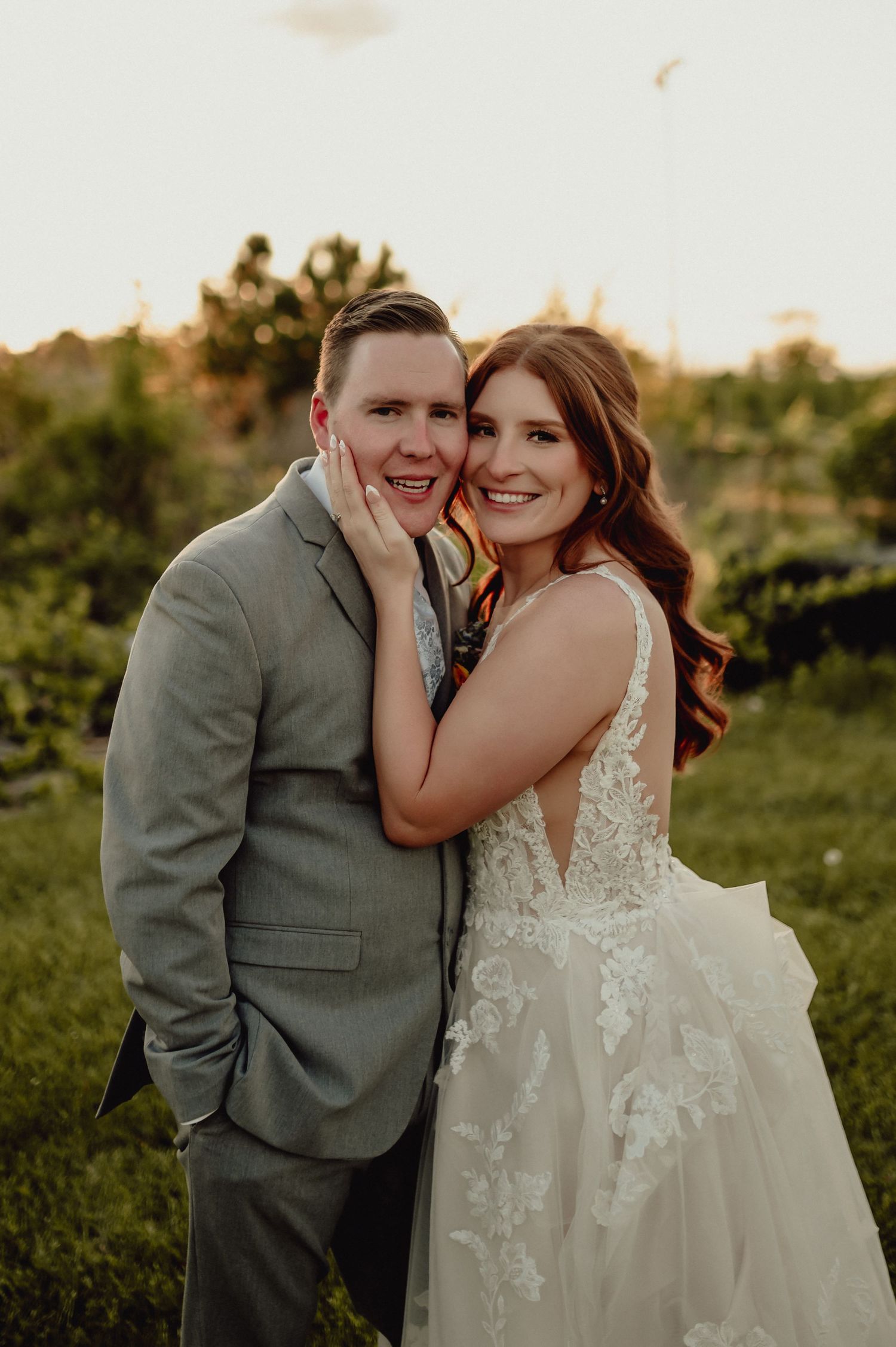 A romantic wedding couple embraces in a golden sunset field wearing a lace gown and gray suit.