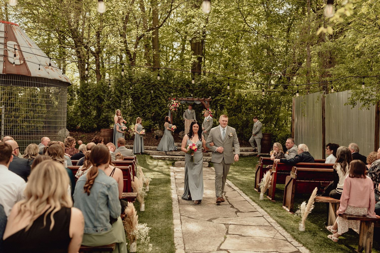 A panoramic view of people gathering in an outdoor garden area with trees and string lights during a social event.