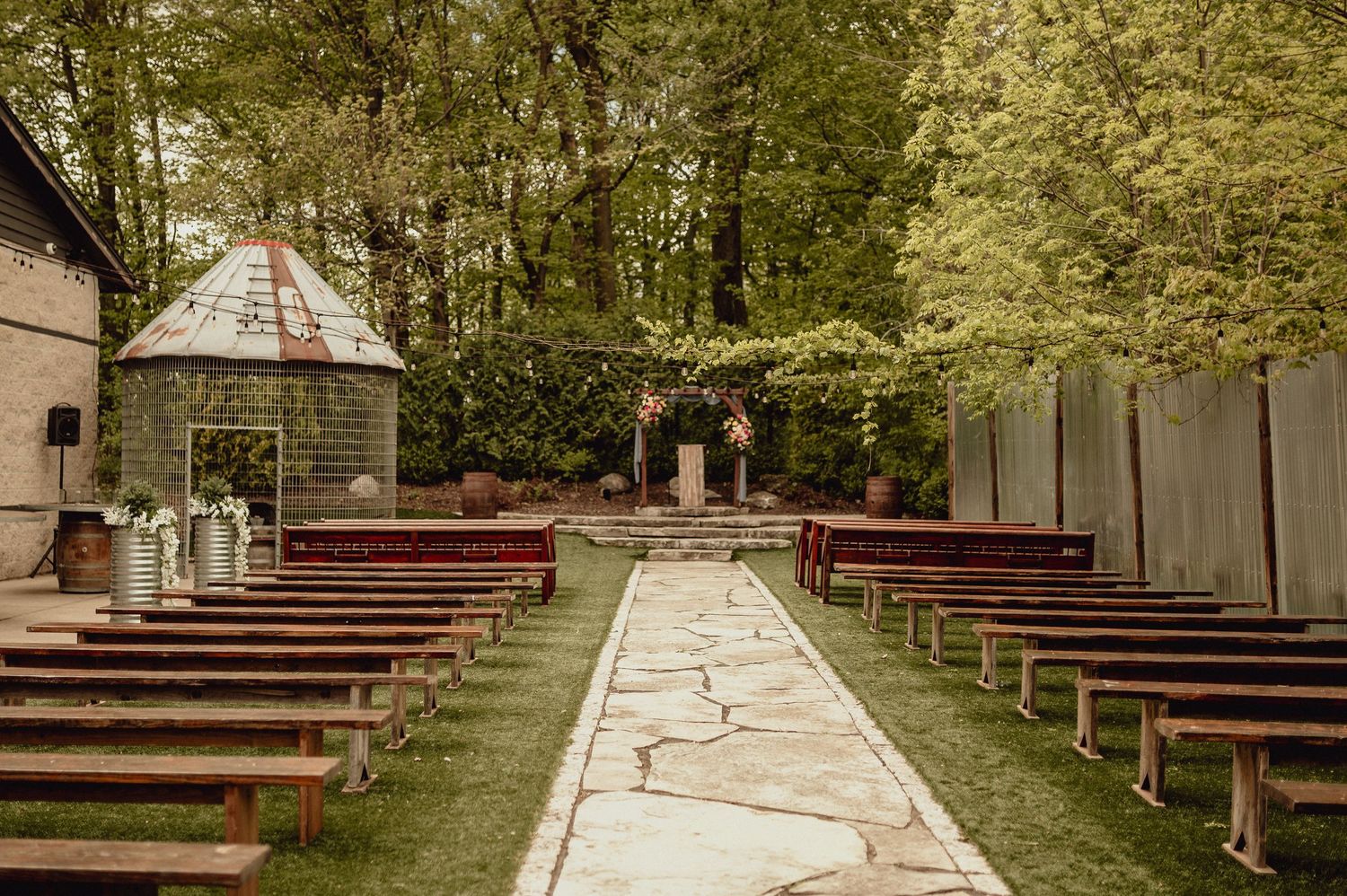 A rustic outdoor wedding venue with wooden benches arranged in rows along a stone pathway surrounded by trees.