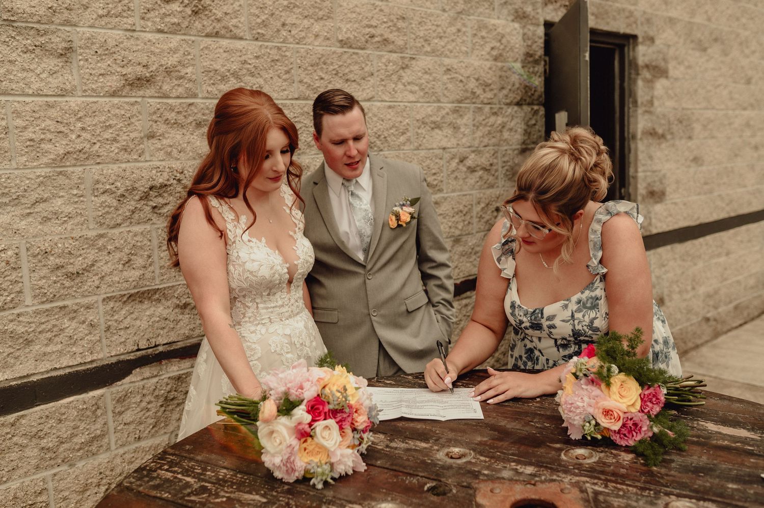 Three people gather around a wooden table signing documents with pink and peach floral bouquets nearby.