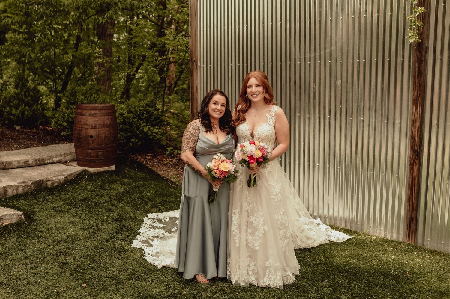 Two people in wedding attire stand together holding bouquets of flowers in an outdoor garden setting.