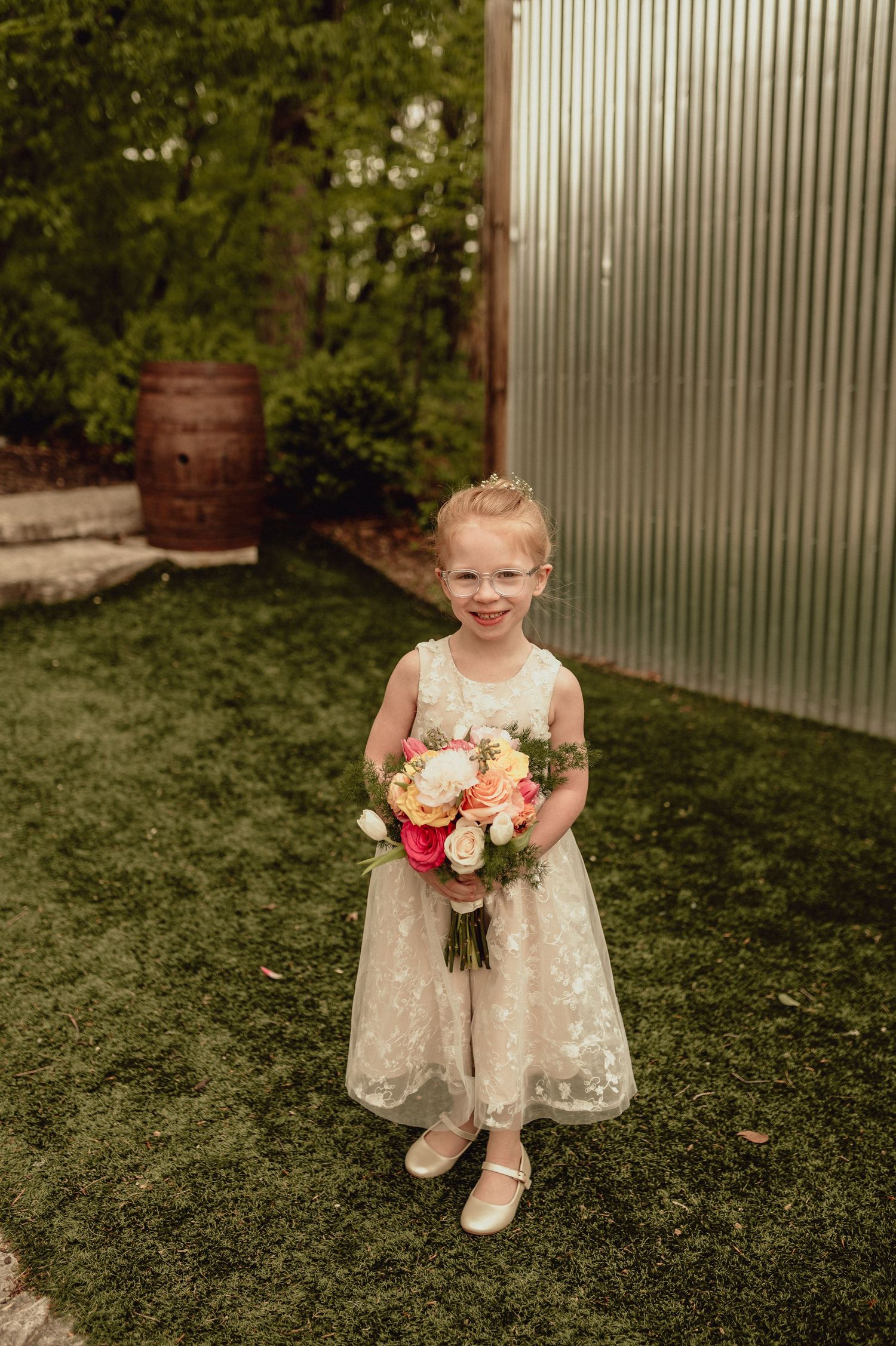 A young flower girl in a white lace dress sits on grass holding a bouquet of pink and peach roses at a wedding.