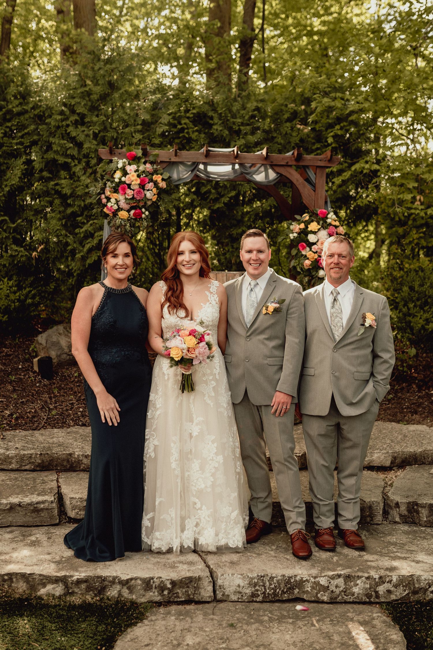 Wedding party members pose together on stone wall in garden setting during outdoor celebration.