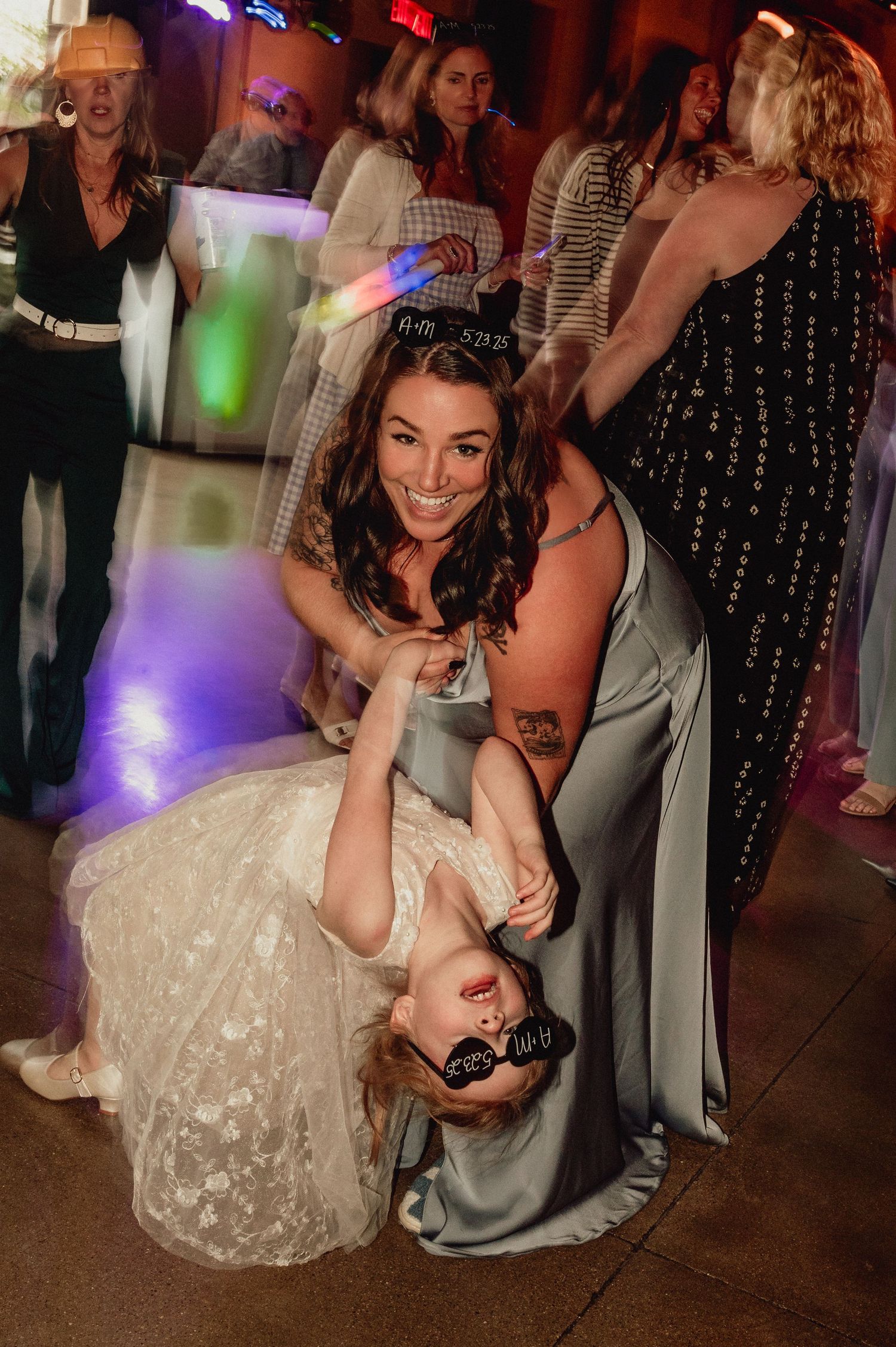 A bride in a white lace dress dips down while dancing at a wedding reception with colorful party lights in the background.