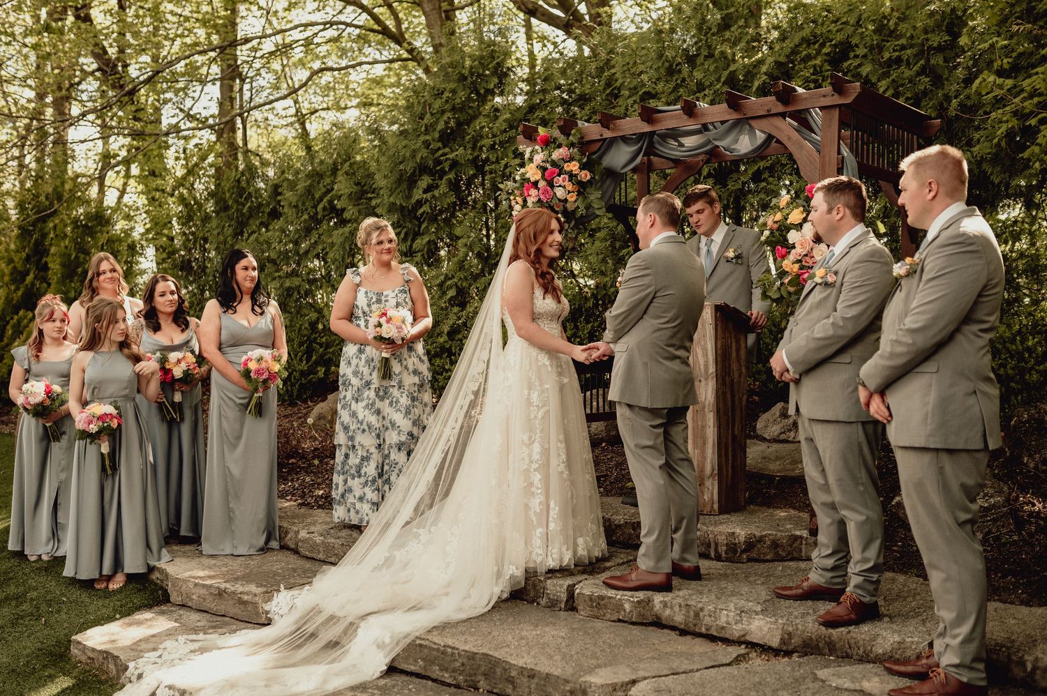 A wedding party stands together outdoors during a ceremony with bridesmaids in gray dresses and groomsmen in gray suits.