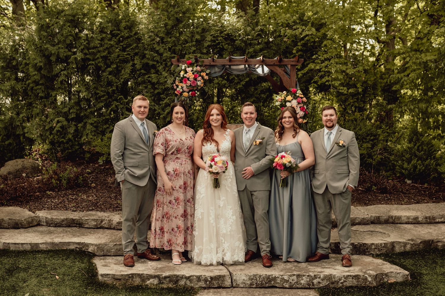 Wedding party wearing pink and gray attire poses together on stone wall in outdoor garden setting.