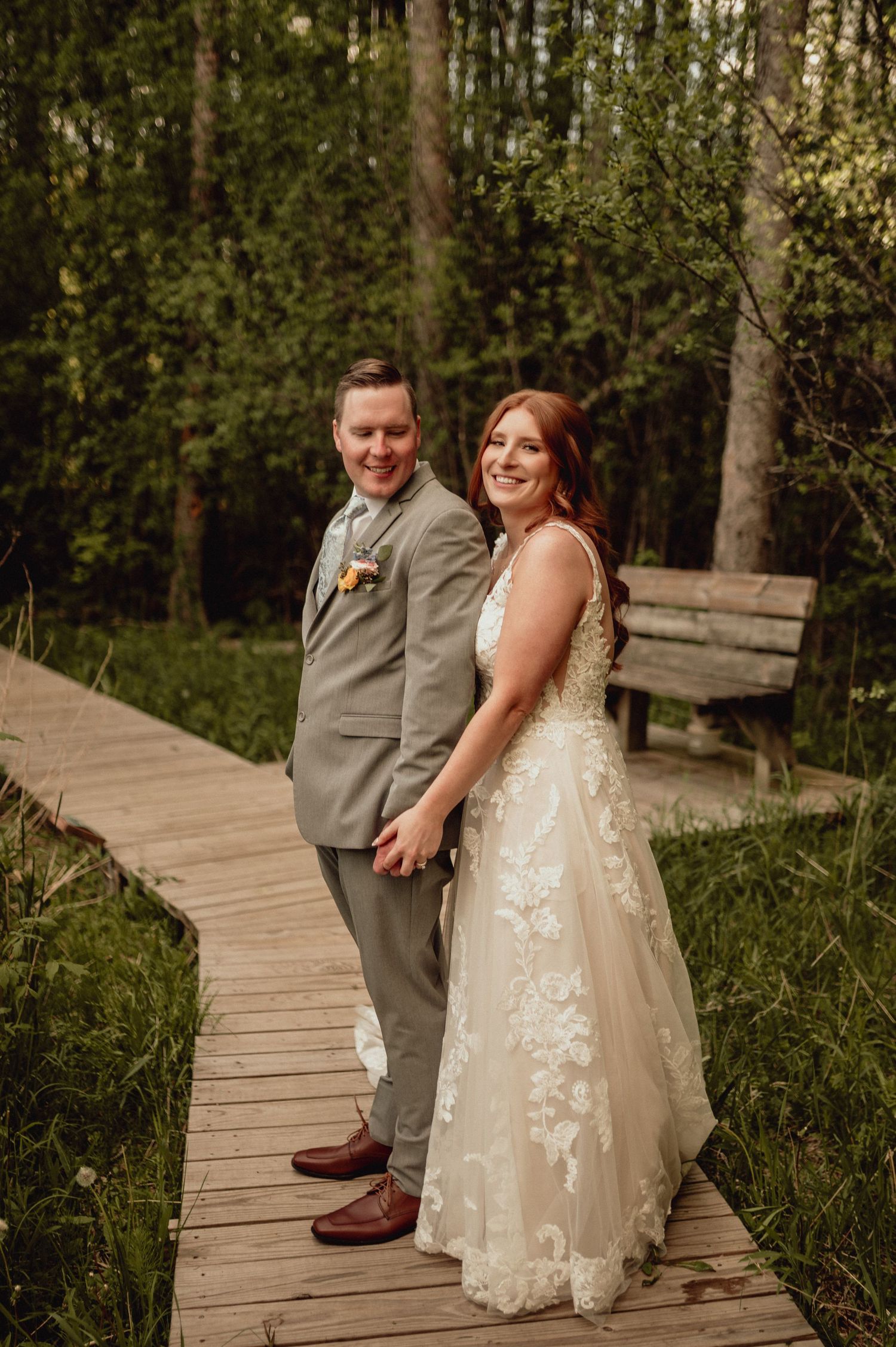 A couple in wedding attire poses on a wooden boardwalk surrounded by lush greenery in a natural outdoor setting.