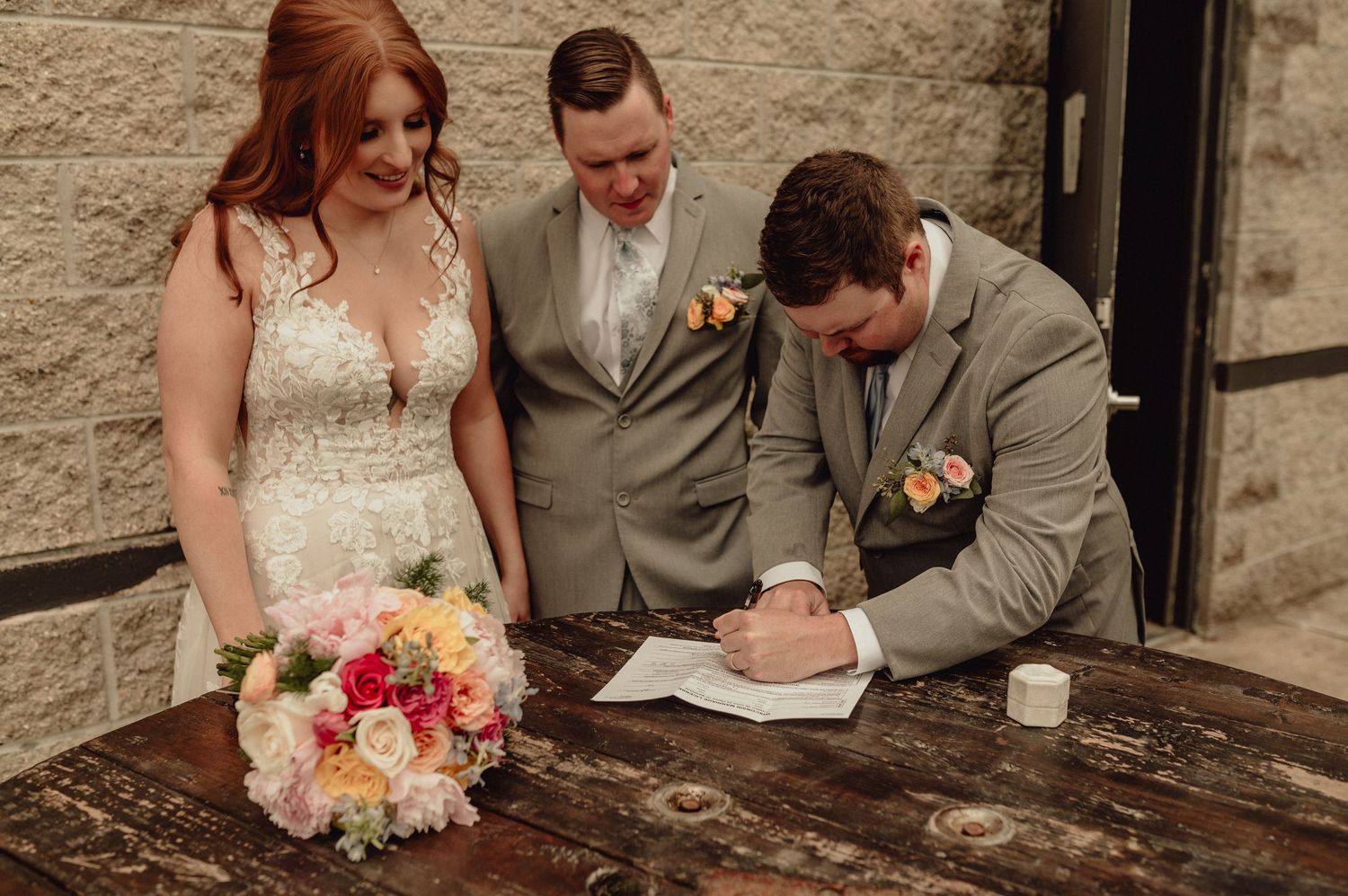 A couple signs their marriage documents at a rustic wooden table with a colorful floral bouquet nearby.
