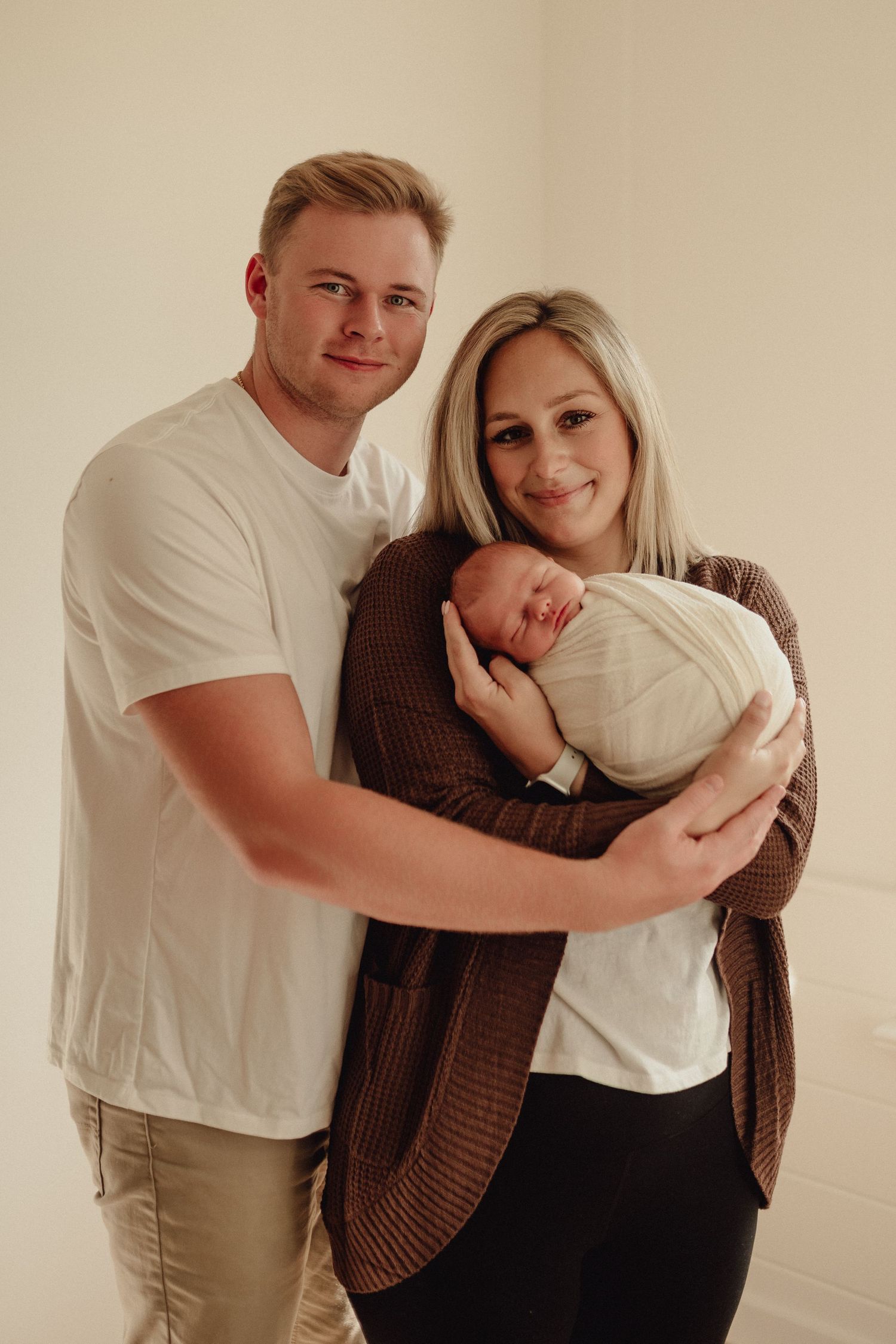 A couple embracing while holding their newborn baby against a neutral wall background.